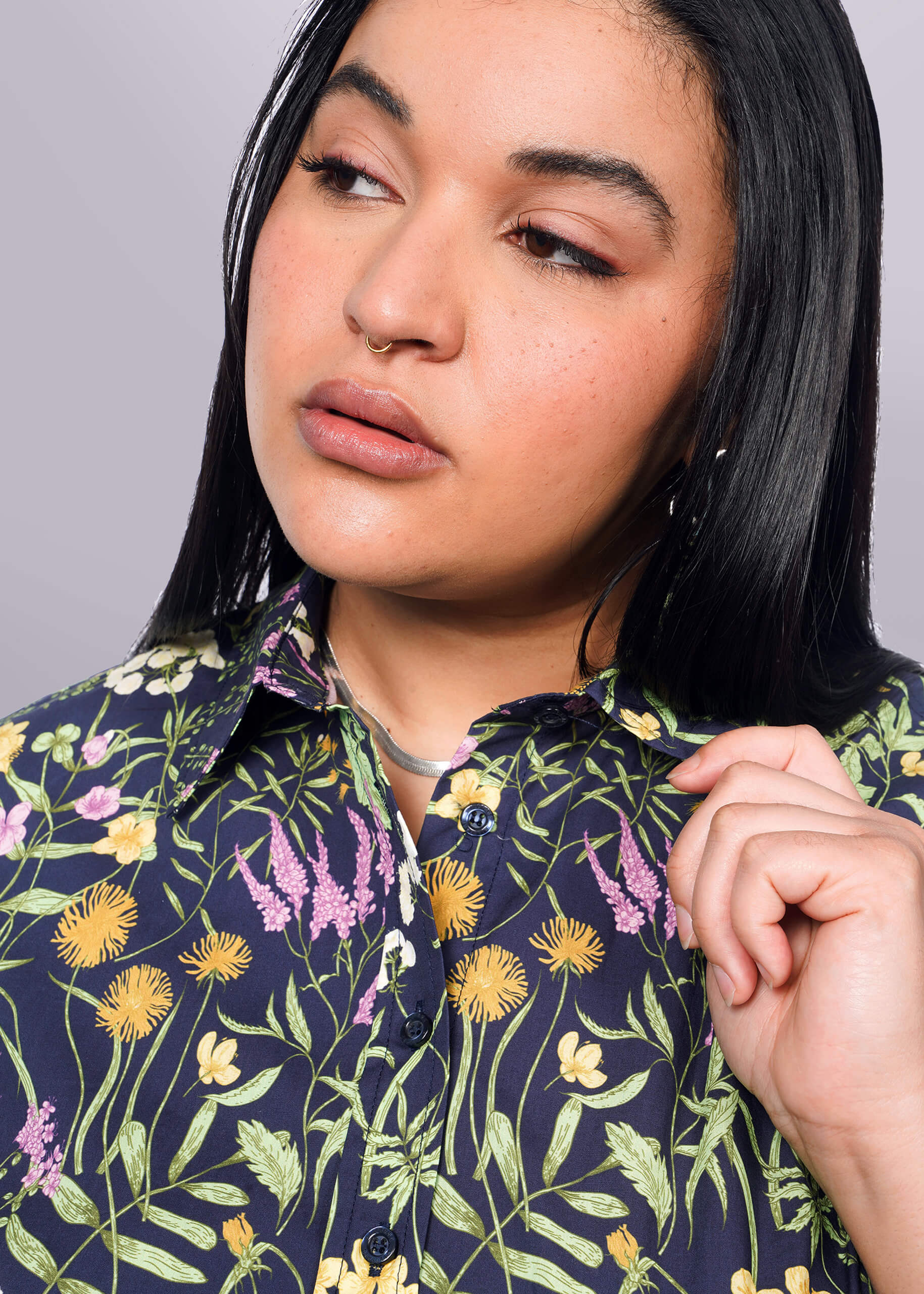 1X= Close up of person with long black hair holding collar of short sleeve button up shirt with botanical print.