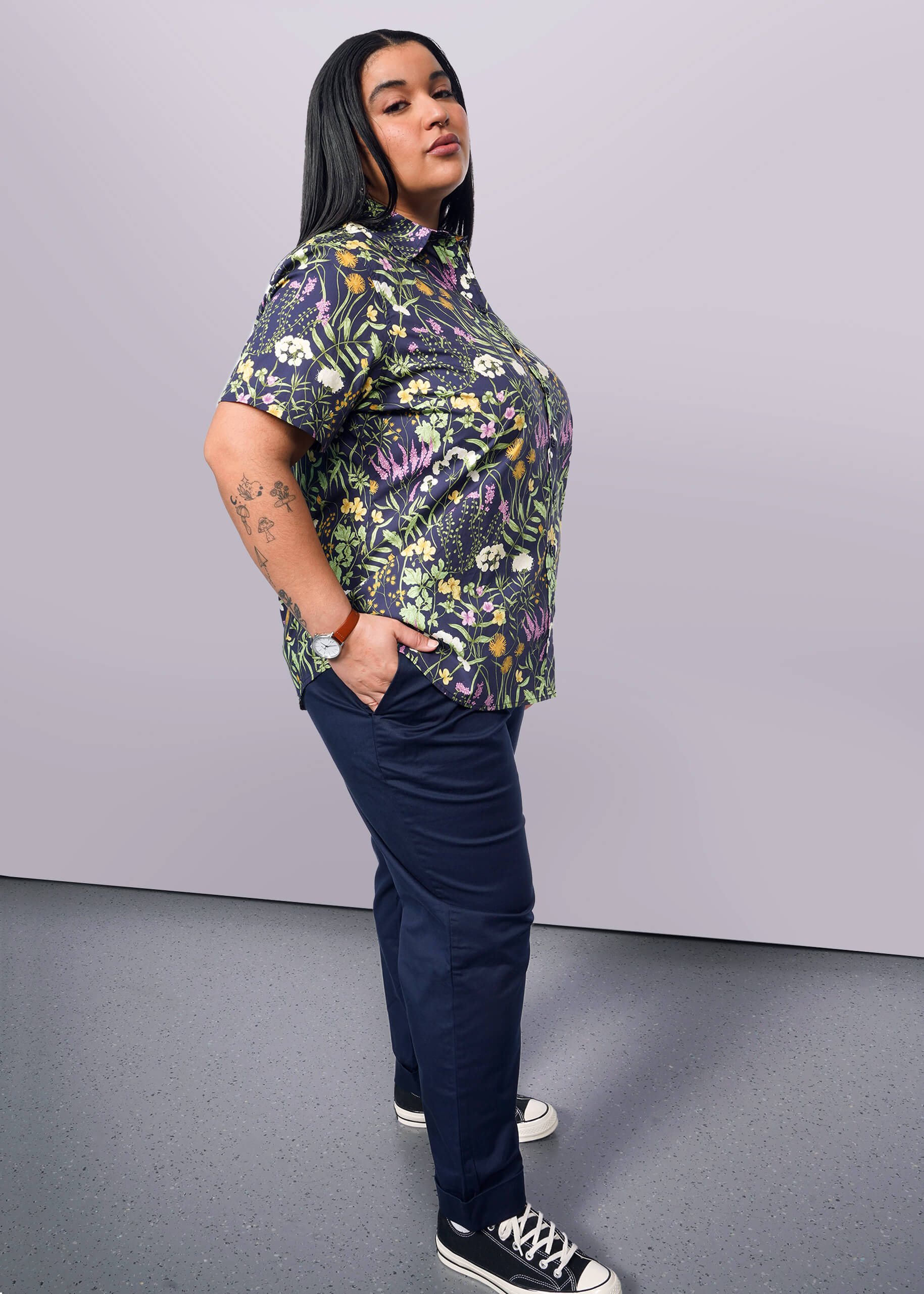 1X=Side view of person with long black hair looking at camera with hand in pant pocket wearing navy trouser, Converse sneakers, and short sleeve button up shirt with botanical print.