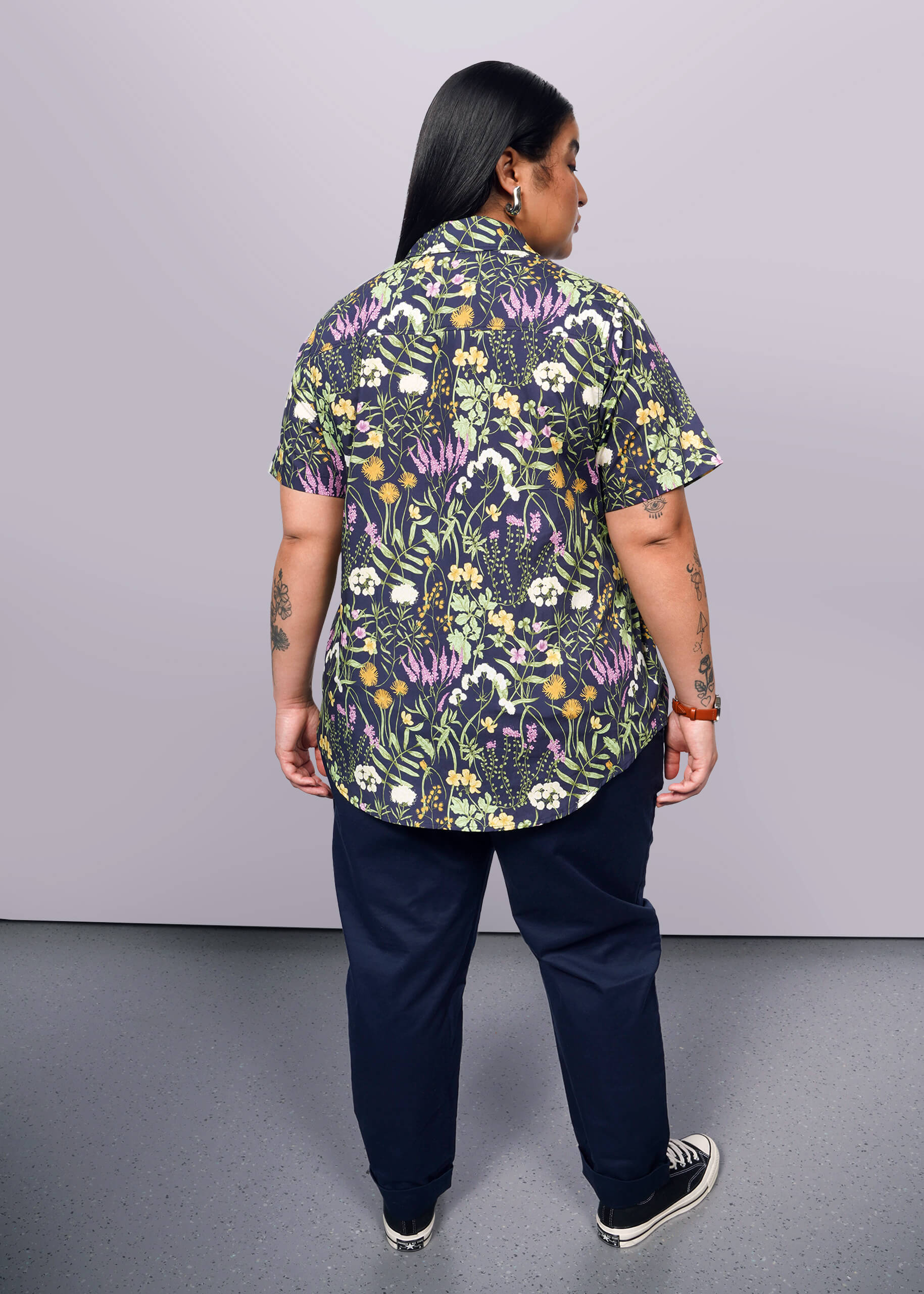 1X=Back view of person with long black hair looking at camera wearing navy trouser, Converse sneakers, and short sleeve button up shirt with botanical print.
