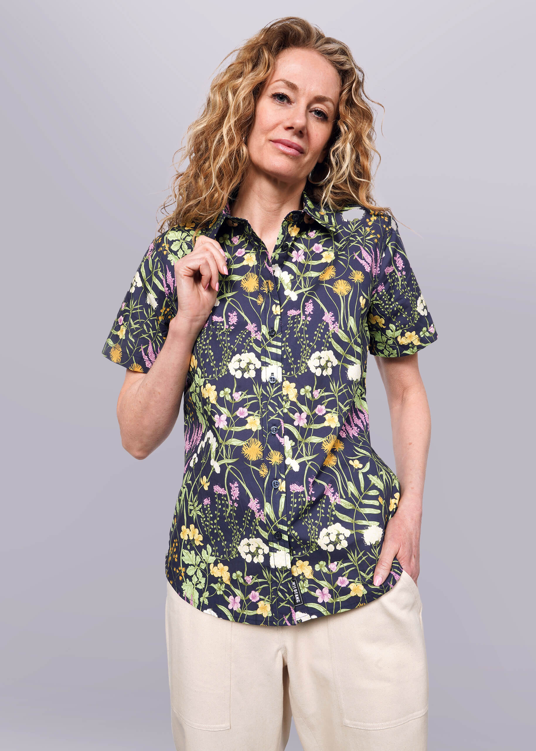 S= Person with wavy blonde hair holds collar of short sleeve botanical printed button up shirt.