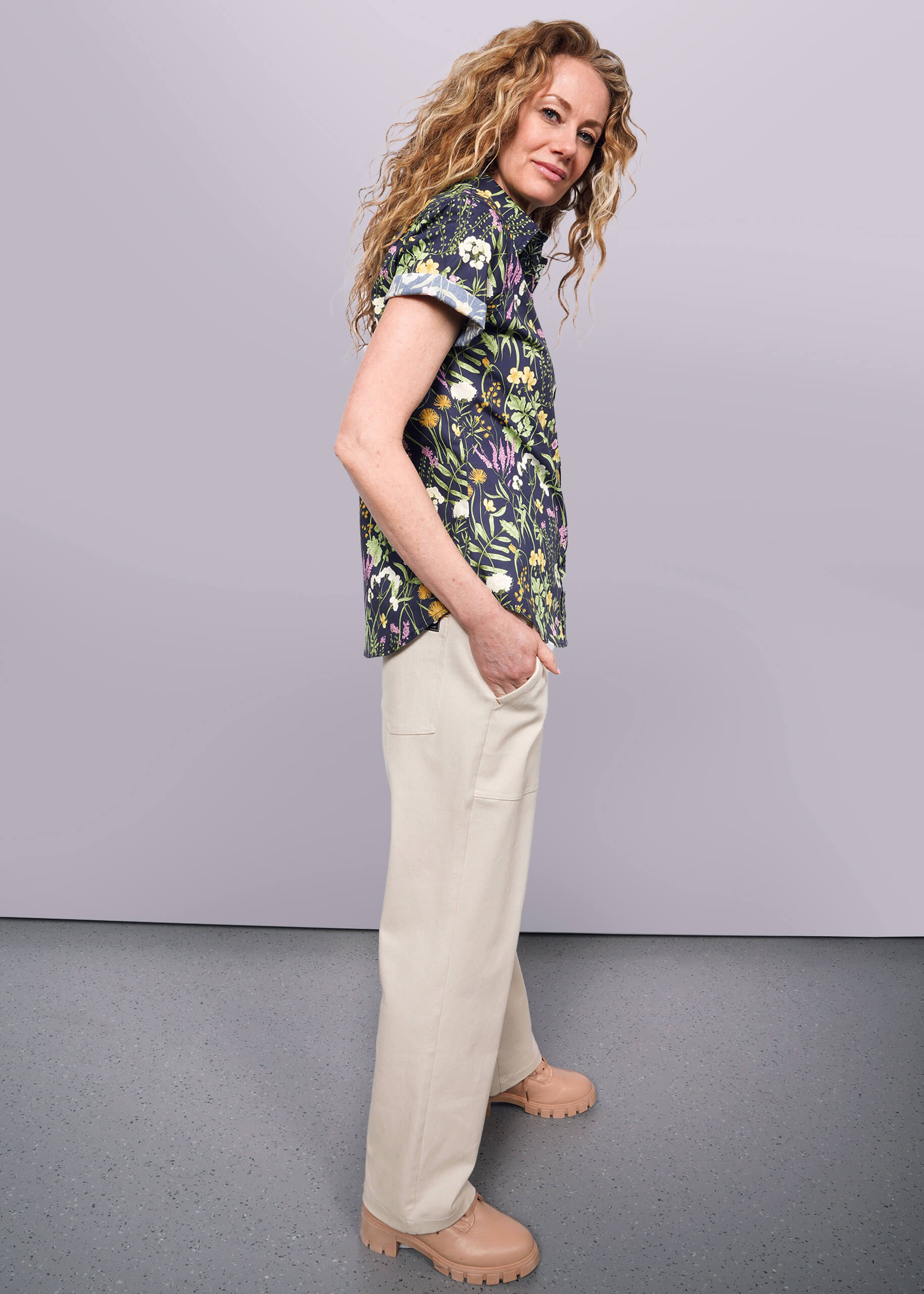 S=Side view of person with wavy blonde hair looking at camera with hand in pocket wearing cream colored utility pant, boots, and short sleeve botanical printed button up shirt.