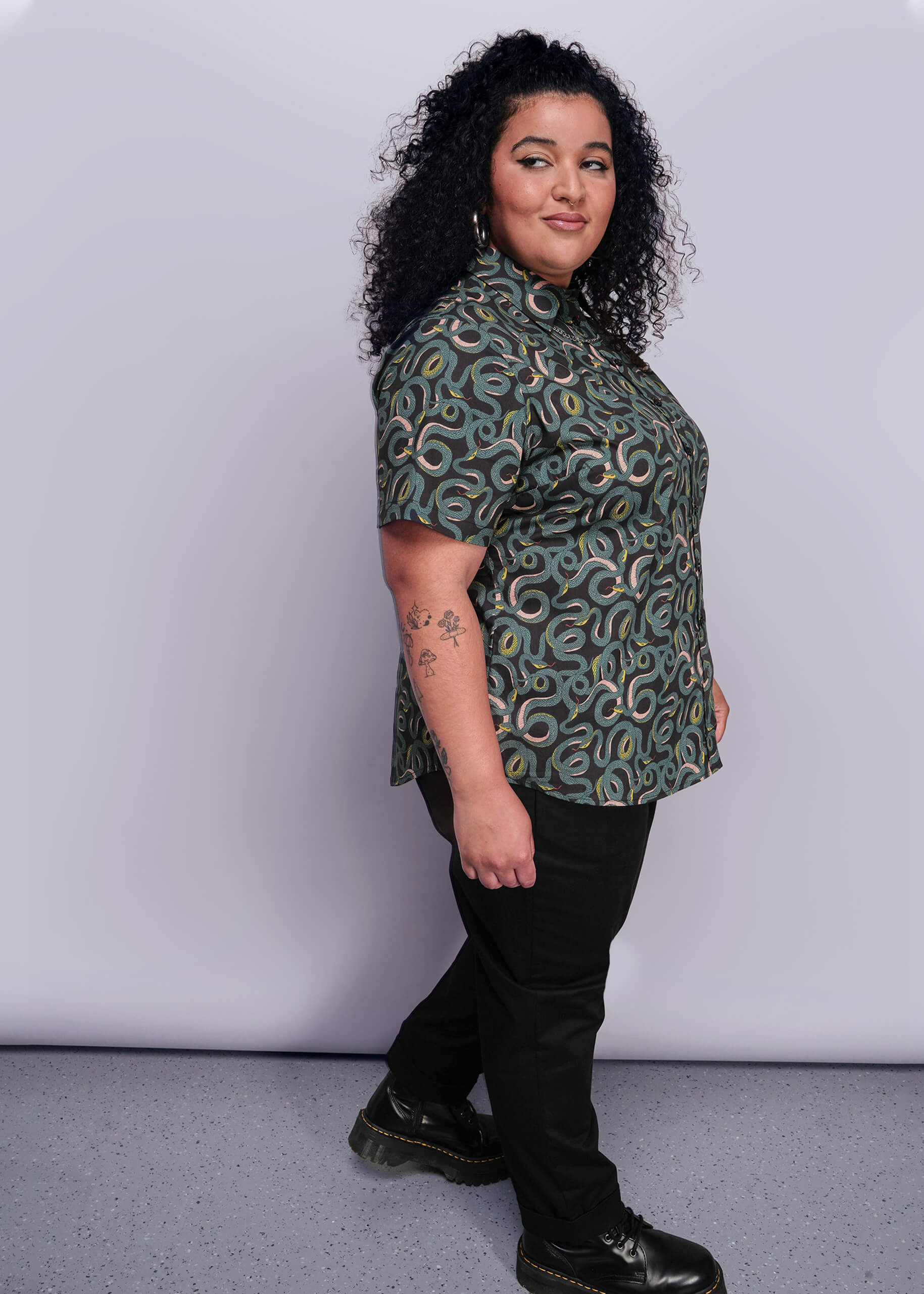 1X=Woman wearing a patterned shirt and black pants against a white background