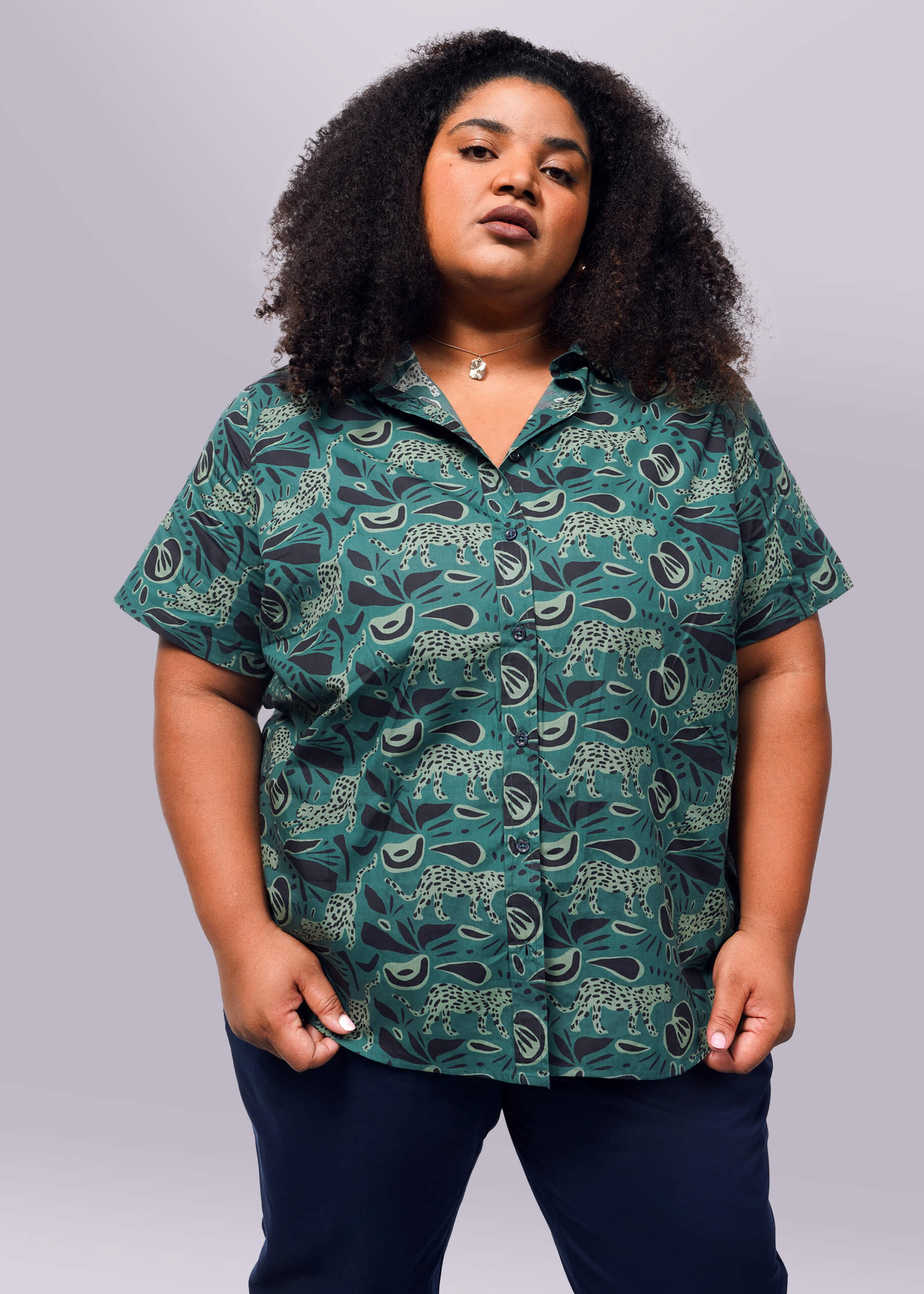 1X= A person with curly hair is wearing the short-sleeved, tailored fit green Wildfang The Essential Button Up shirt featuring a leopard print and black pants. This breathable cotton outfit stands out against a plain white background.