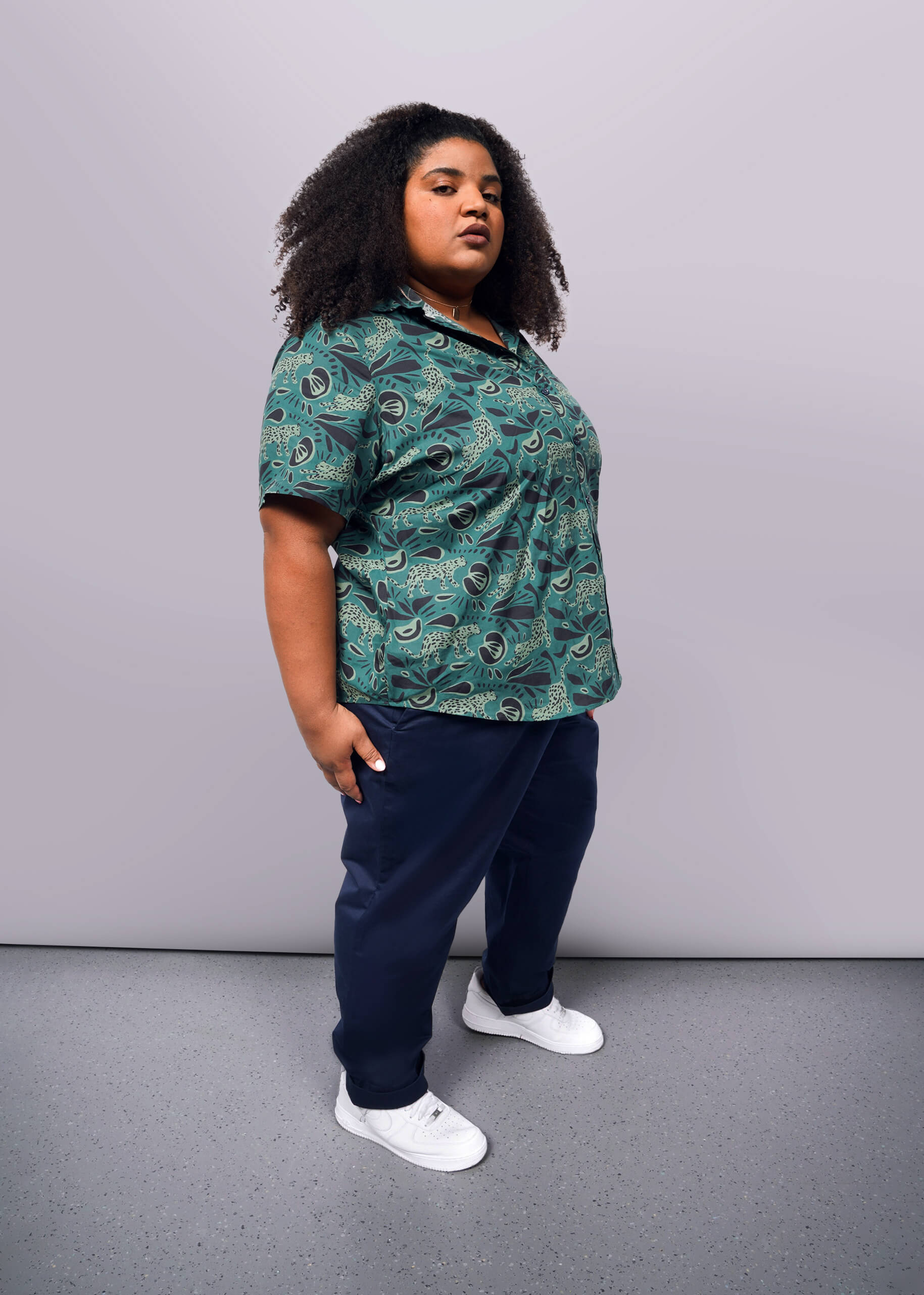 1X= Wearing The Essential Button Up and navy patterned pants, complemented by white sneakers, a person stands confidently. Their long curly hair frames a determined expression against a light background, while breathable cotton fabric ensures comfort in their tailored fit ensemble.