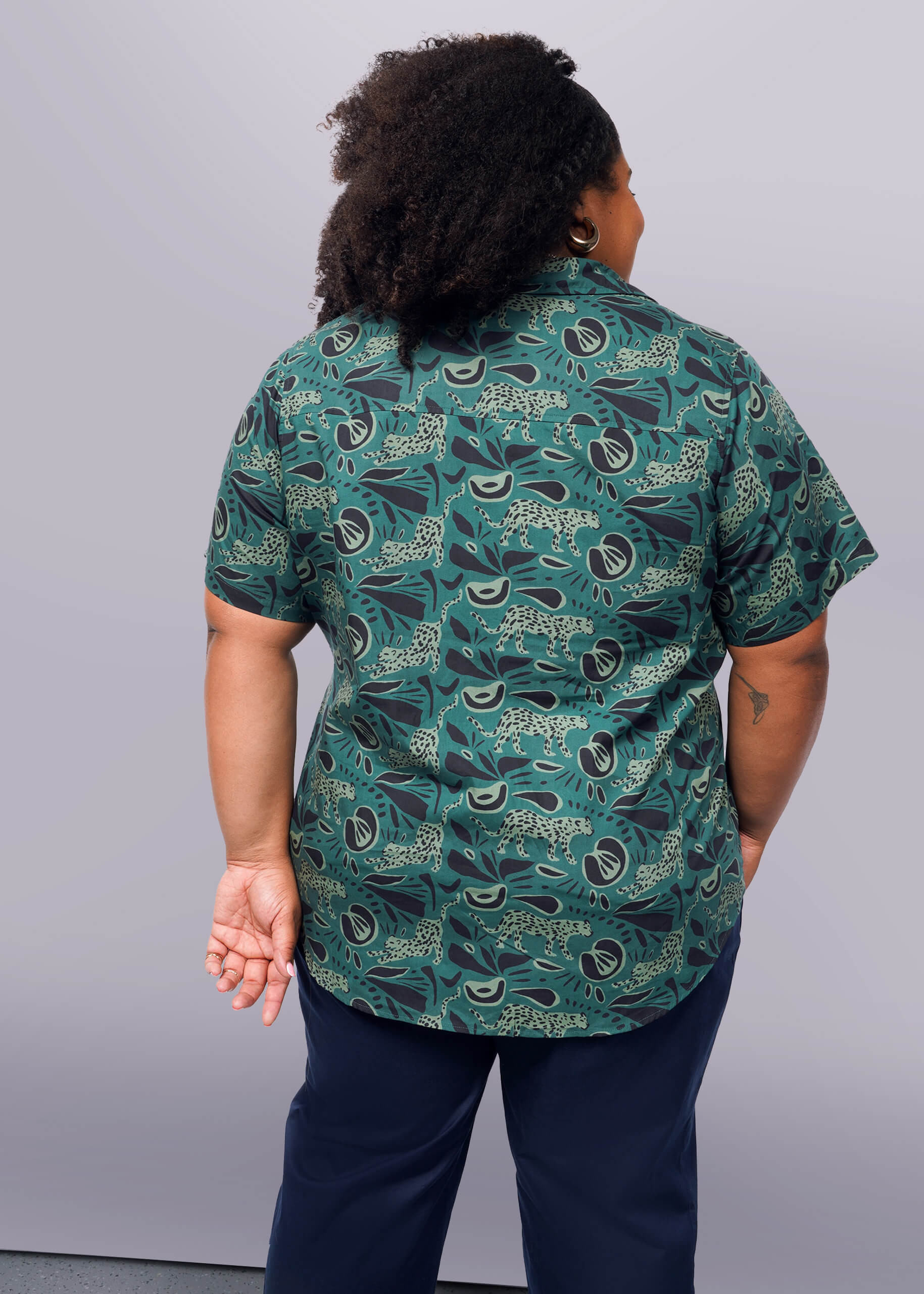 1X=Someone with curly hair faces away, sporting The Essential Button Up, a green patterned Wildfang shirt made from breathable cotton. Dark pants complement the tailored-fit top as their arms rest relaxed against a plain white wall.