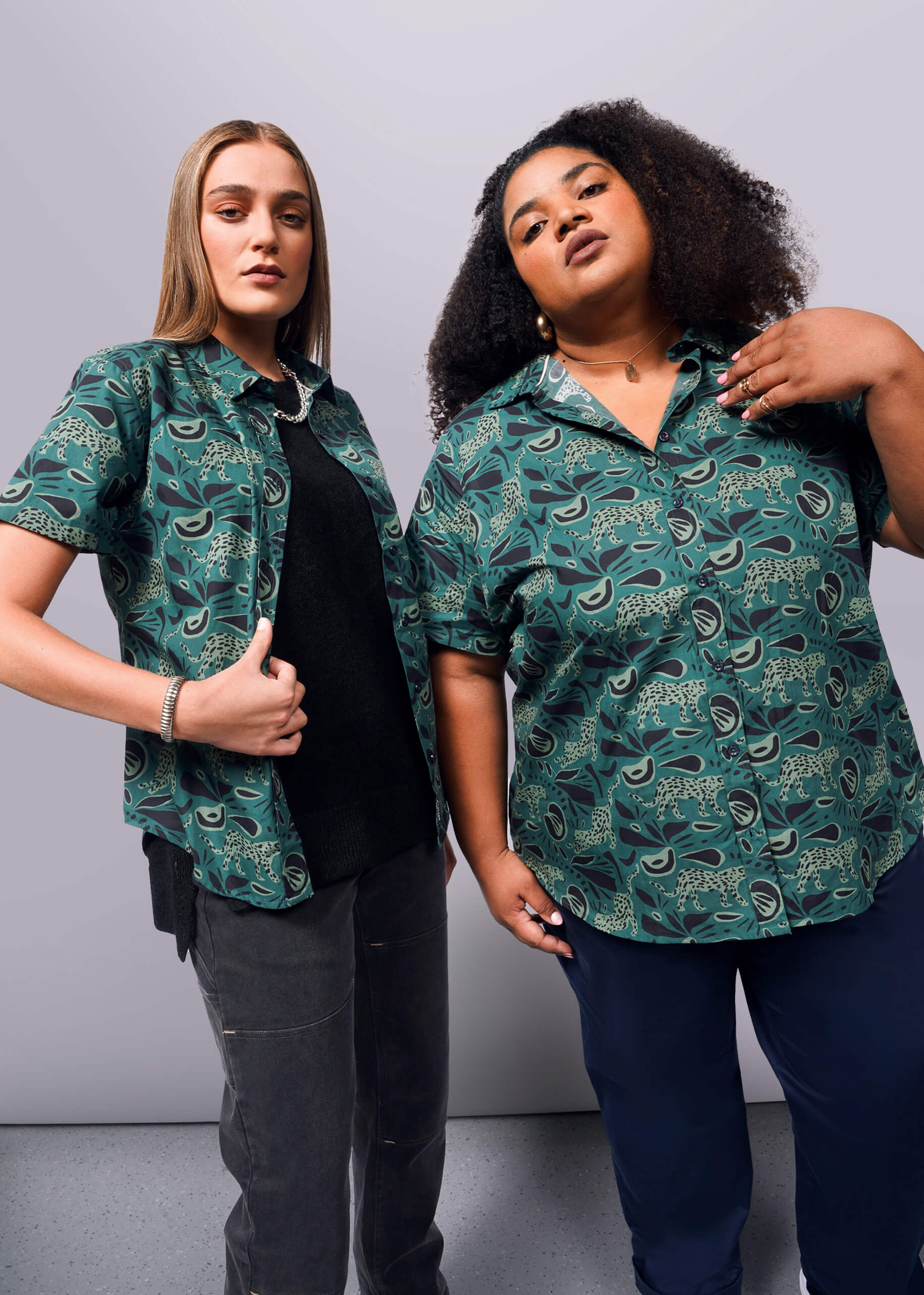 Two people stand confidently against a white background, showcasing The Essential Button Up shirts from Wildfang in classic green pattern. Made of breathable cotton, the tailored fit enhances their style. One has straight hair and dark pants, while the other has curly hair and navy pants.
