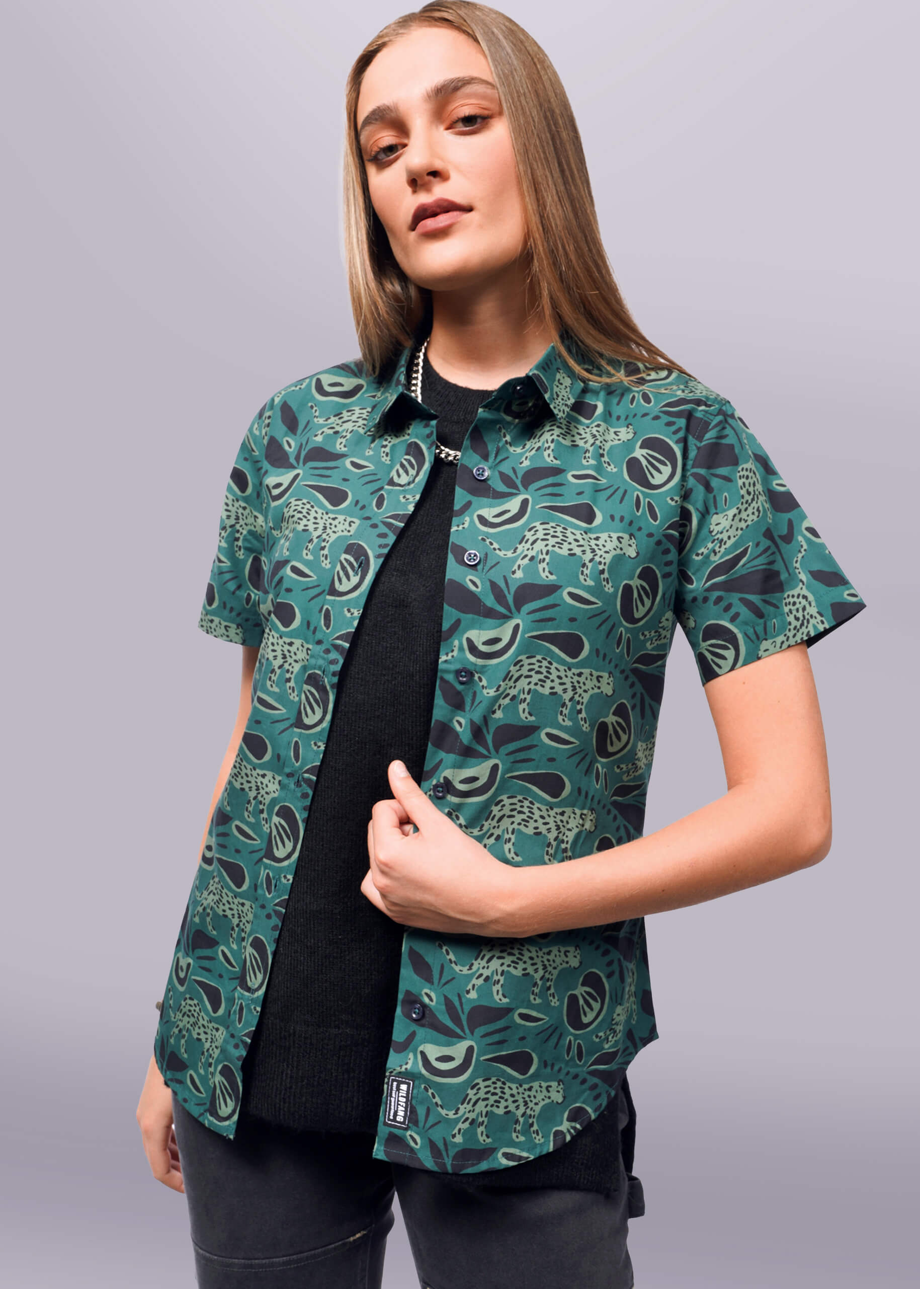 S= A person with long hair confidently poses in The Essential Button Up by Wildfang, featuring a dark green leopard and leaf pattern over a black top. Made from breathable cotton, the shirt offers a tailored fit. They maintain a neutral expression against a plain white background.