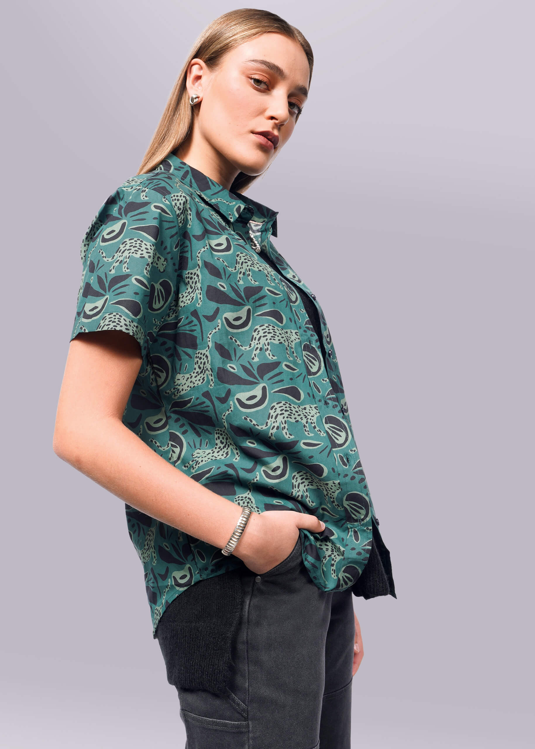 S=A person with long hair wears The Essential Button Up, tailored in breathable cotton with short sleeves, and dark pants. They stand confidently to the side, one hand in a pocket, against a white background.