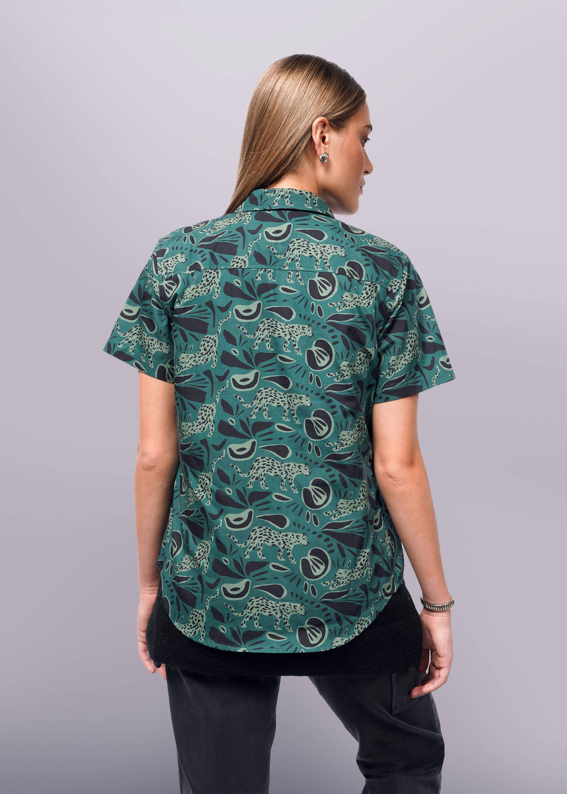 S=A person with long hair, wearing a green The Essential Button Up by Wildfang, features a black and white abstract pattern. Facing slightly to the side in dark pants and a bracelet, they stand against a plain white background.