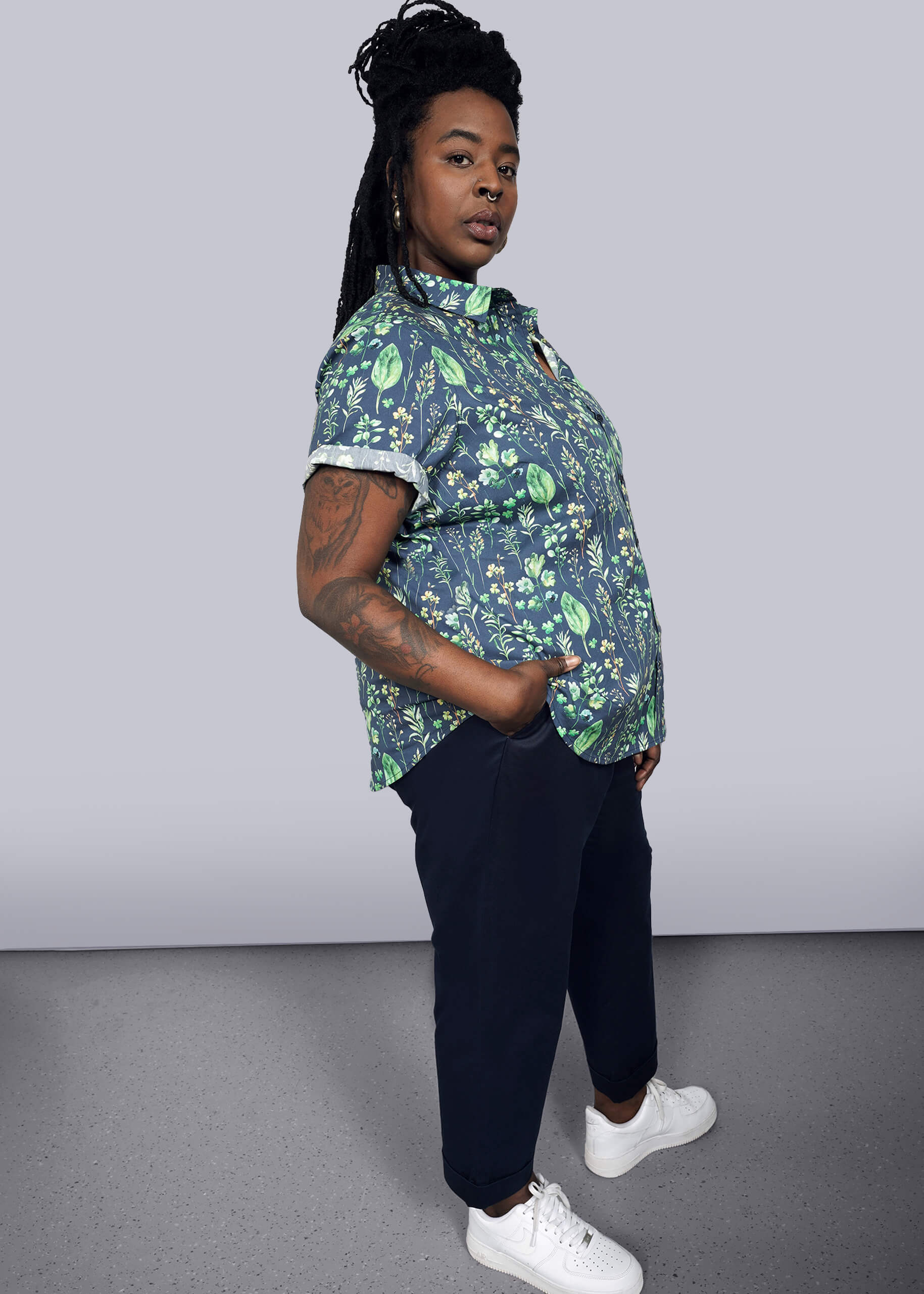 1X= With long braided hair, the person stands confidently with one hand in their pocket, wearing The Essential Button Up, a tailored fit leaf-patterned shirt with short sleeves, paired with dark pants and white sneakers. Made from 100% cotton, they look assuredly towards the camera.
