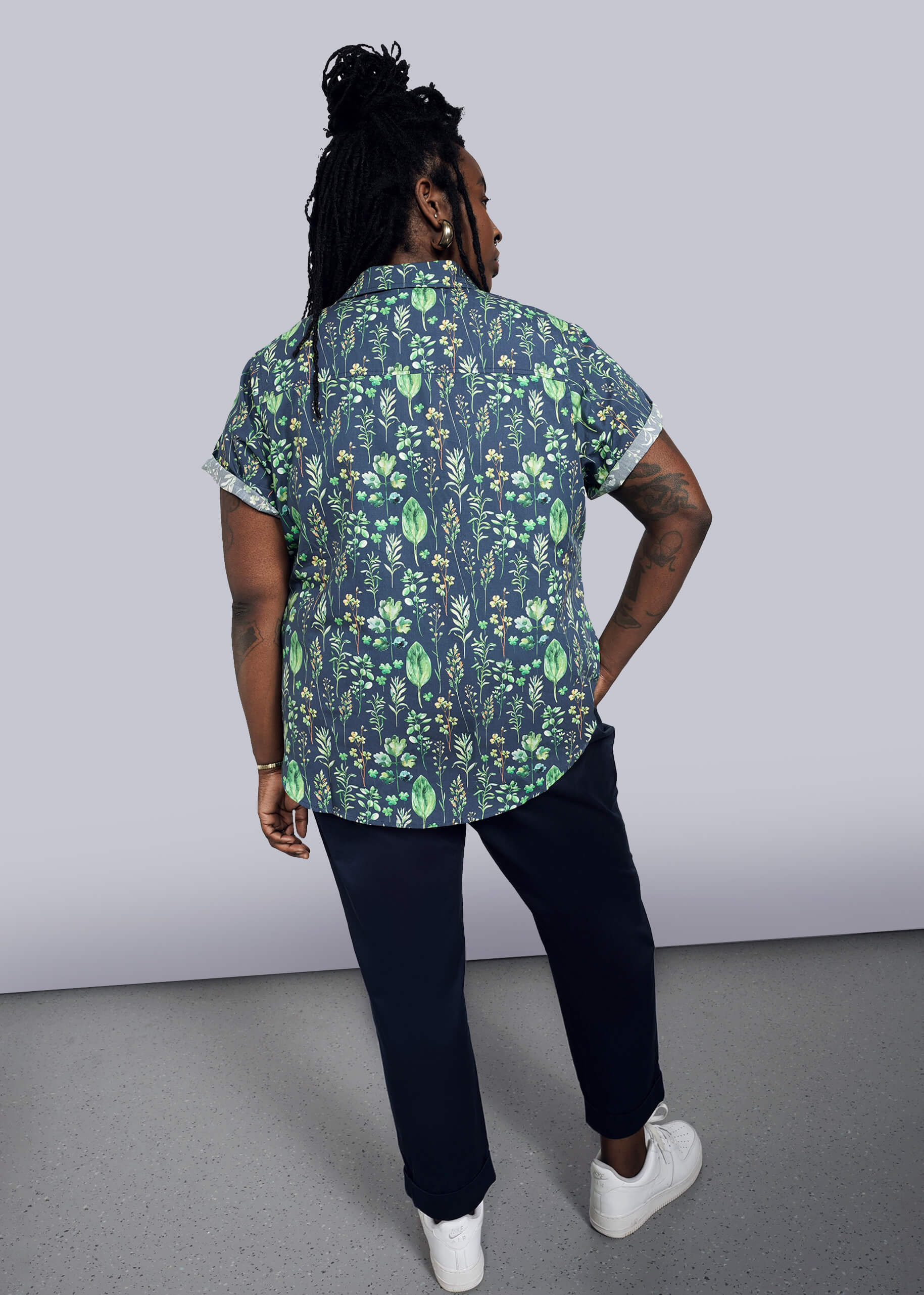 1X=Wearing The Essential Button Up, a short-sleeved, 100% cotton shirt in a tailored fit with a green leaf pattern, the person with long braided hair stands back to the camera. They pair it with dark pants and white sneakers against a plain, minimalistic background.