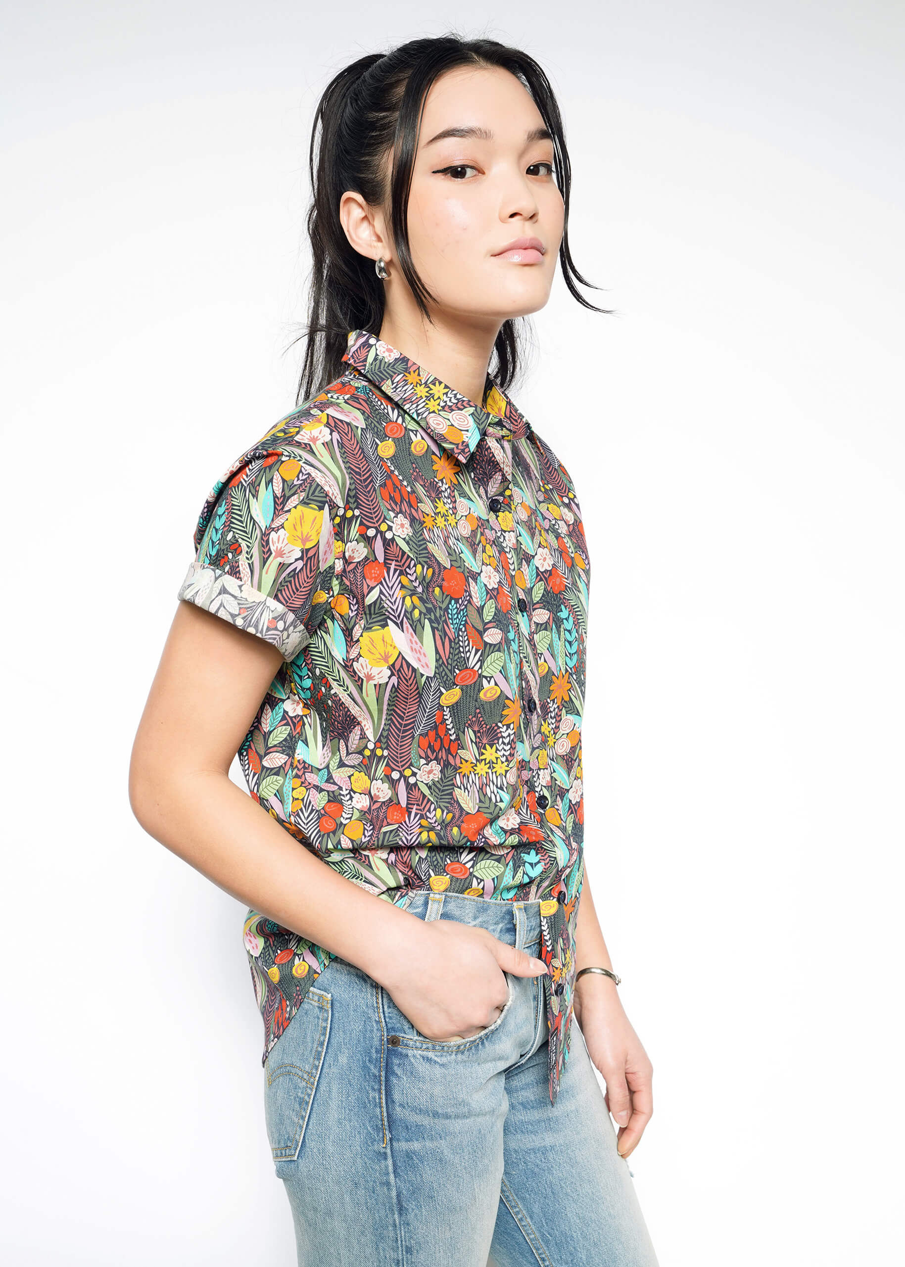 S=A person stands against a light background wearing The Essential Button Up, a colorful patterned short-sleeve shirt in breathable cotton, paired with blue jeans. Their hair is styled in a ponytail with loose strands framing their face, one hand in a pocket, and a neutral expression.