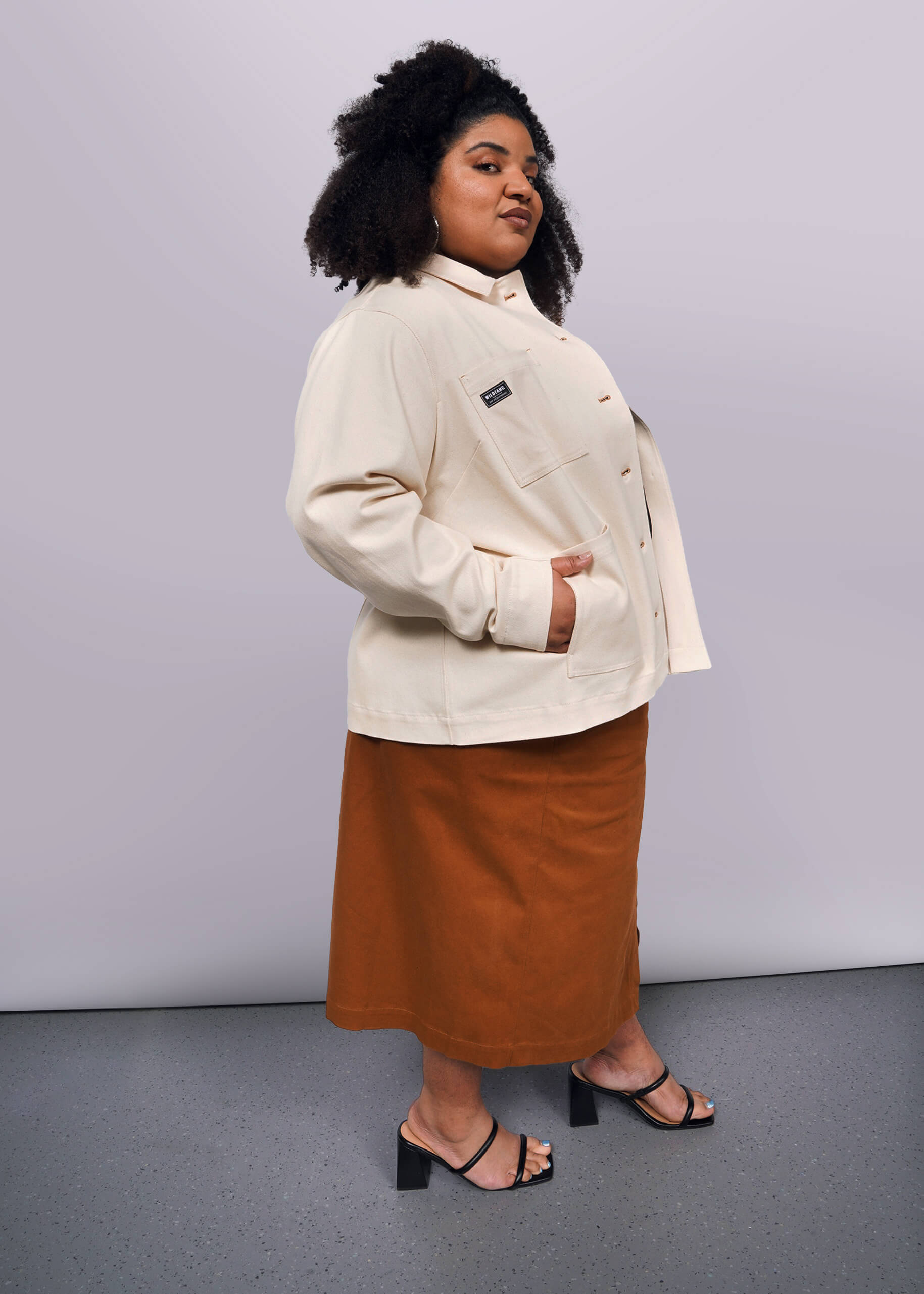 1X=A person with curly hair confidently wears The Essential Denim Utility Jacket with deep pockets, paired with a brown skirt and black heeled sandals, standing in a studio against a neutral background.