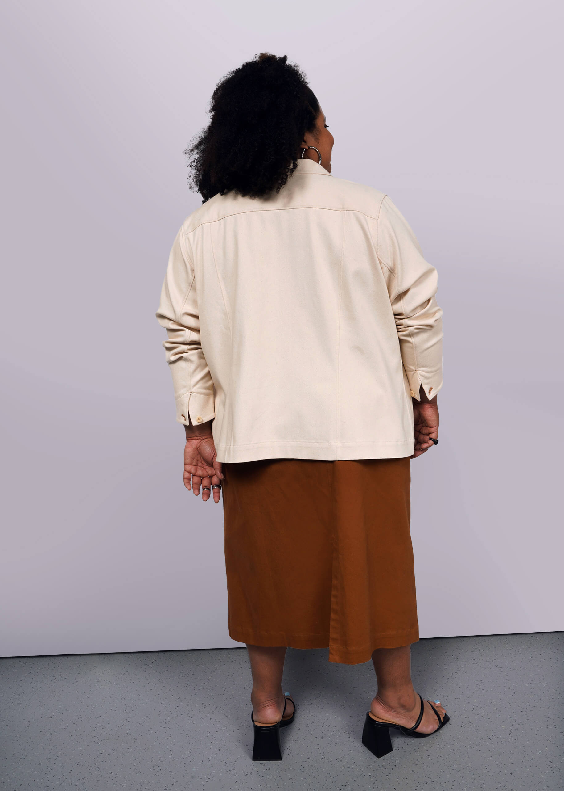 1X=A person with curly hair stands with their back to the camera in a beige long-sleeve shirt, brown skirt, and black open-toe heels. Completing the look is The Essential Denim Utility Jacket, featuring deep pockets. The background is plain white.
