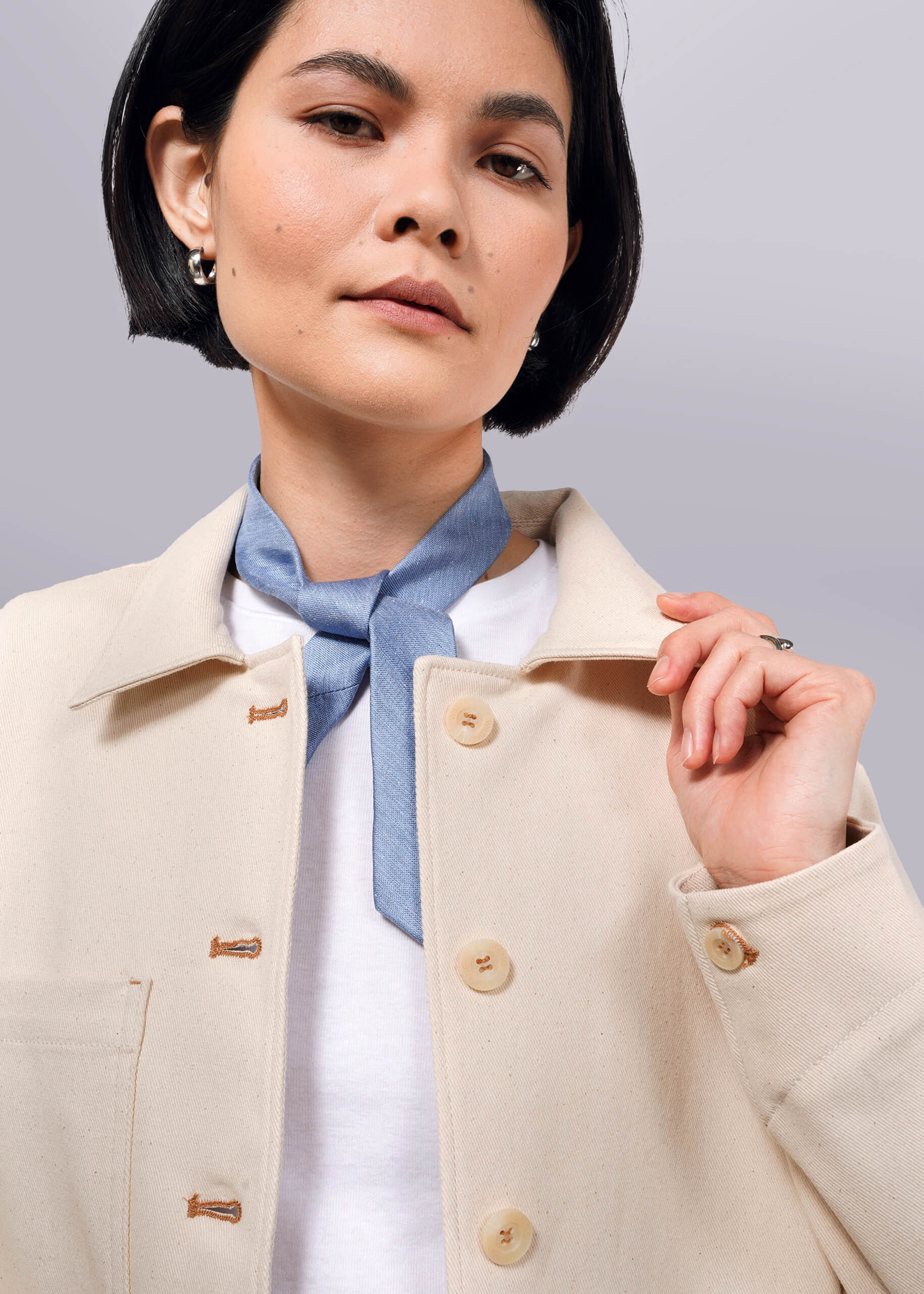 S=A woman with short black hair wears The Essential Denim Utility Jacket, featuring large buttons and deep pockets. She accessorizes with a blue neck scarf and small hoop earrings, holding the collar and looking slightly upward against a white background.