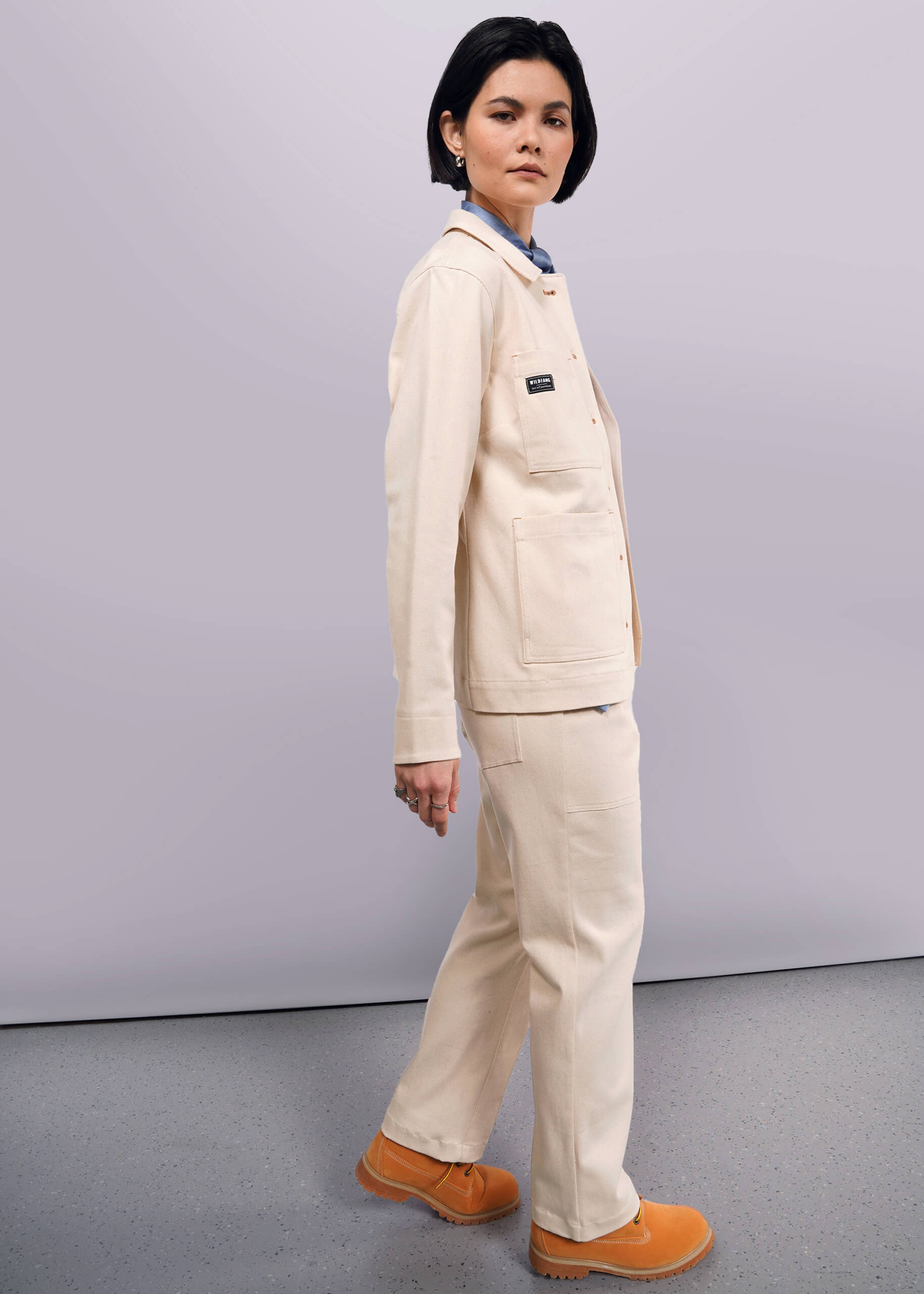 S=A person with short dark hair wearing The Essential Denim Utility Jacket paired with matching pants and tan shoes stands on a gray speckled floor against a light background. Deep pockets on the beige jacket combine style and stretch comfort as they look sideways.