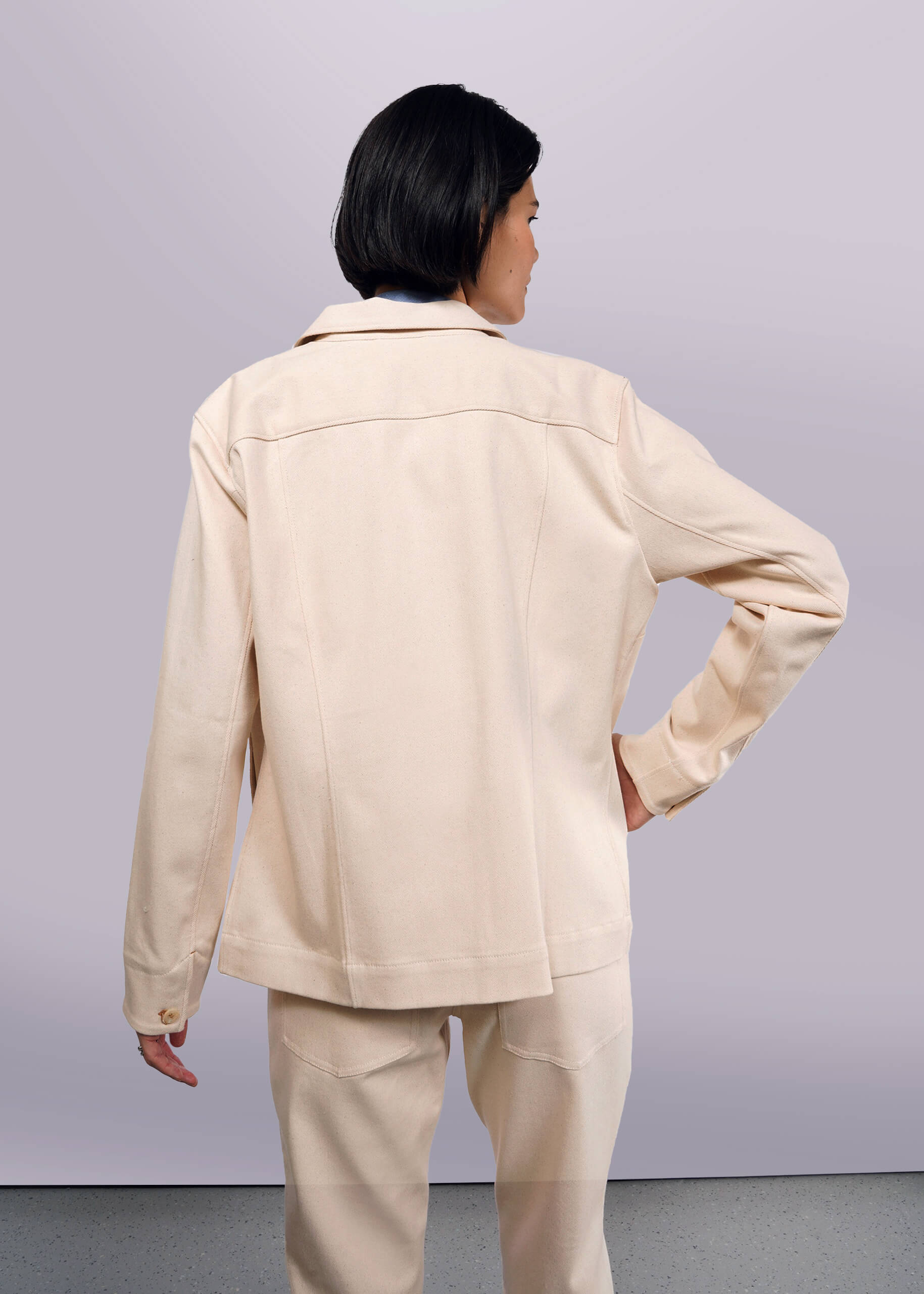 S=A person with short dark hair stands back to the camera wearing The Essential Denim Utility Jacket and matching pants in light beige. The outfit offers stretch comfort for all-day wear, highlighted against a plain light gray background, enhancing the sleek design of the ensemble.