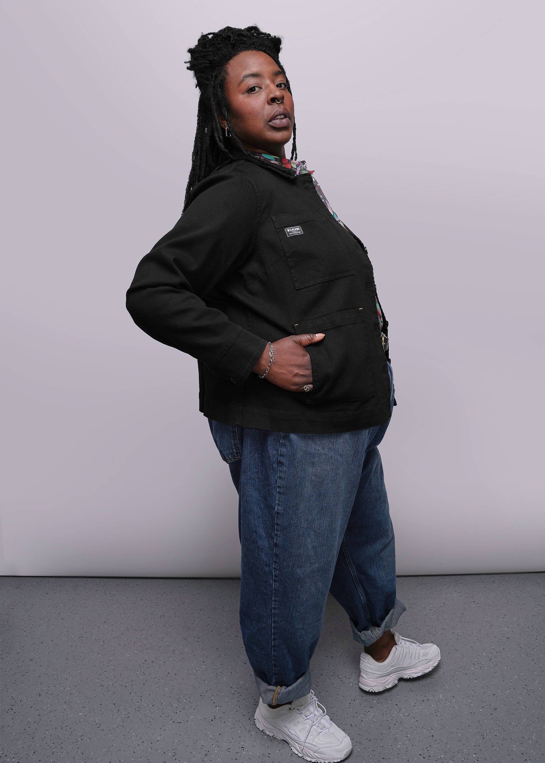 1X=Person wearing a black denim jacket, blue jeans, and white sneakers against a plain background