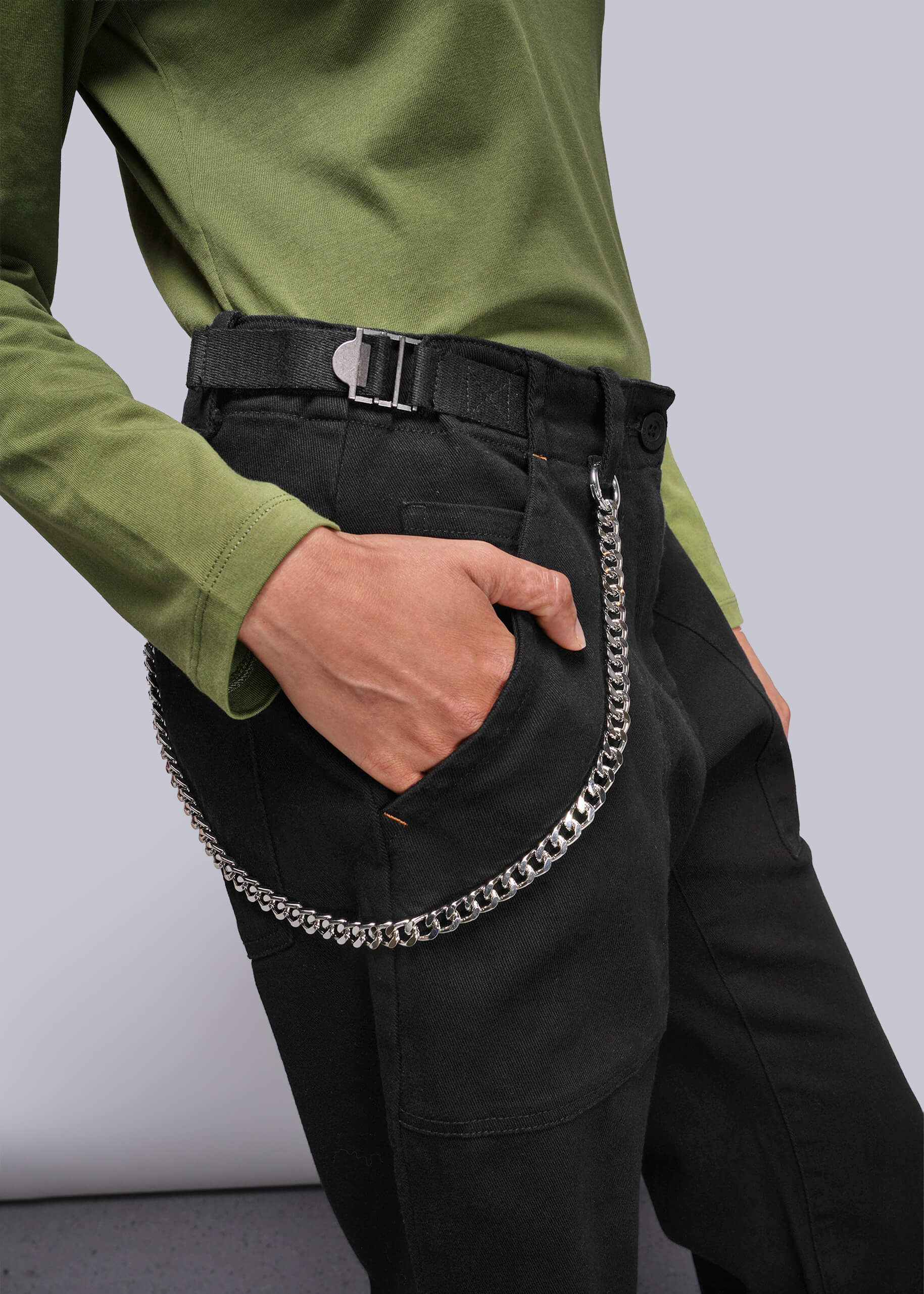 S=Person wearing a green shirt and black pants with a chain detail, standing against a plain background.