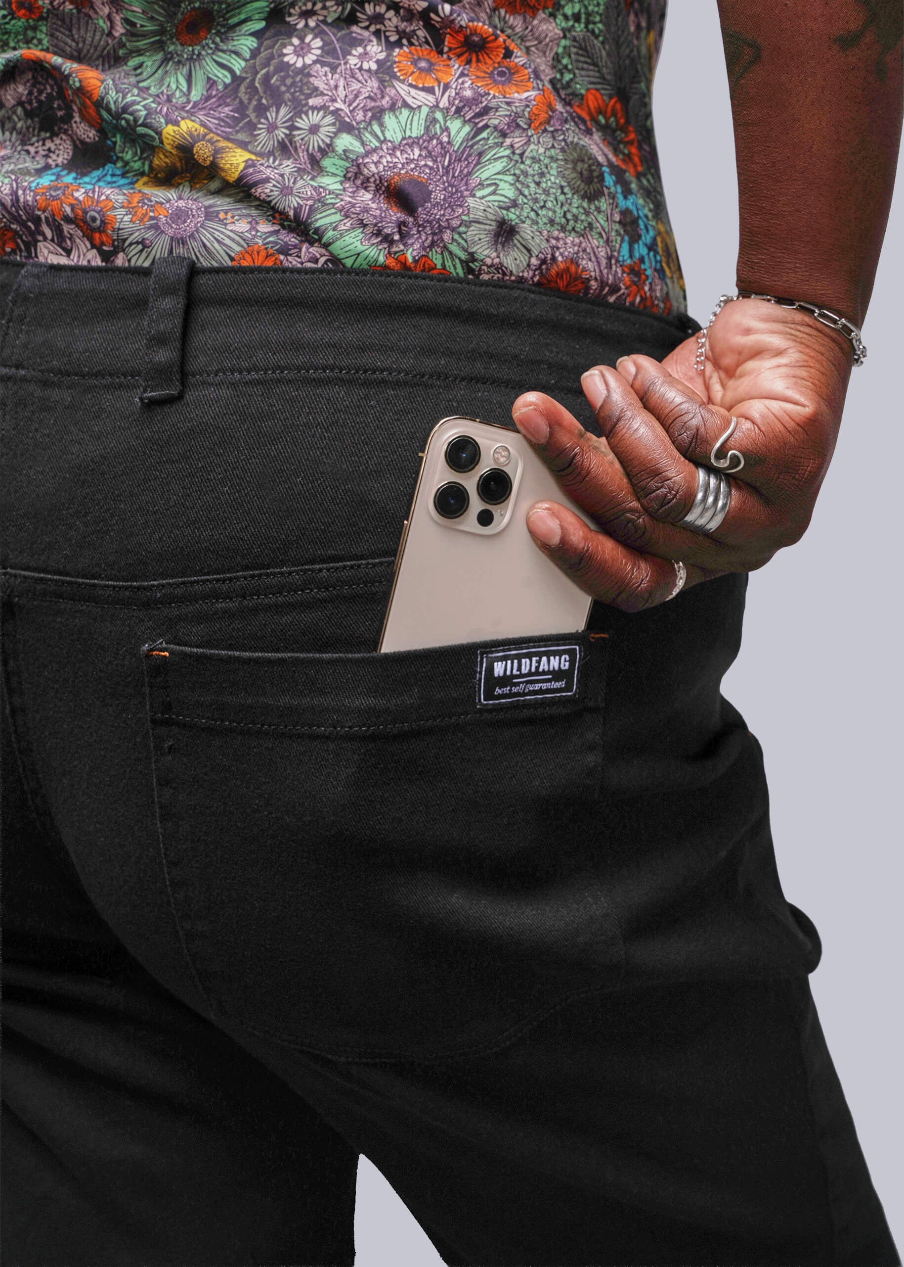 1X=Close up of person wearing black pants with a Wildfang label, putting phone in pocket, against a neutral background