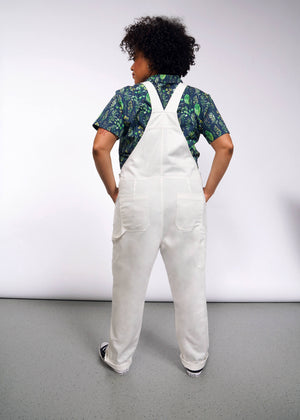 1X=A person with curly hair facing away wears The Essential Herringbone Overall over a blue and green patterned shirt. On a gray speckled floor against a gray wall, their hands are tucked into the overalls deep pockets.
