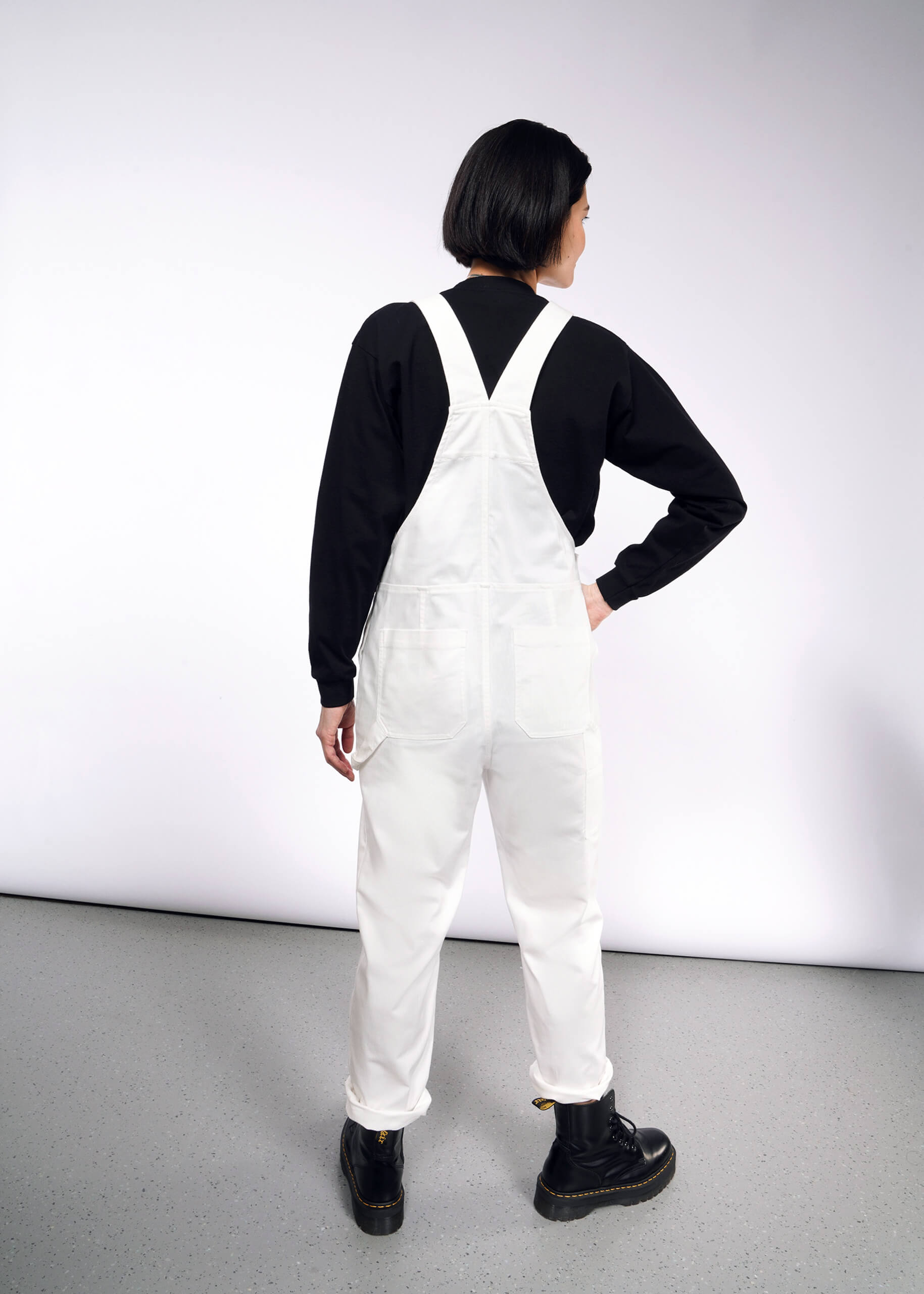 S= A person with short black hair in The Essential Herringbone Overall, adjustable straps, and a black long-sleeve shirt stands hand on hip in a white room with a gray floor, wearing black boots. They face away from the camera.