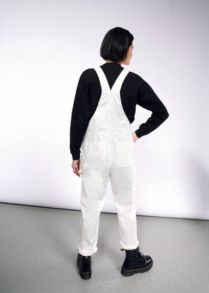 S= A person with short black hair in The Essential Herringbone Overall, adjustable straps, and a black long-sleeve shirt stands hand on hip in a white room with a gray floor, wearing black boots. They face away from the camera.