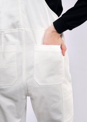 S=A person wearing The Essential Herringbone Overall in white cotton with a black top has their hand in the deep back pocket, showcasing the fabrics texture and stitching against a plain background, highlighting the lower back.