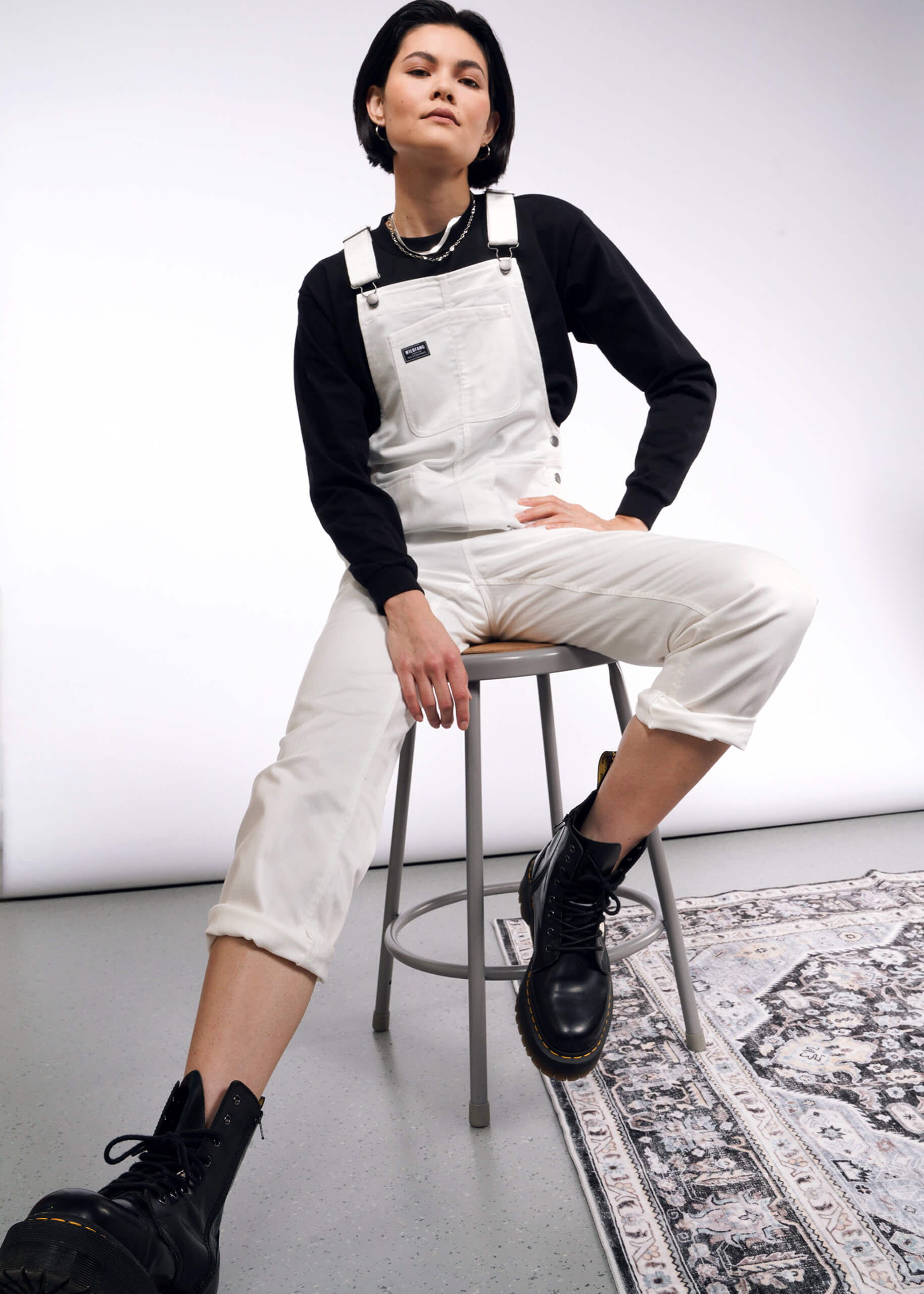 S=A person confidently sits on a stool wearing the Essential Herringbone Overall with adjustable straps, a black long-sleeve shirt, and sturdy black boots. Their stylish pose on a patterned rug beautifully contrasts with the simple white and gray backdrop.