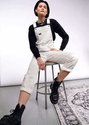 S=A person confidently sits on a stool wearing the Essential Herringbone Overall with adjustable straps, a black long-sleeve shirt, and sturdy black boots. Their stylish pose on a patterned rug beautifully contrasts with the simple white and gray backdrop.