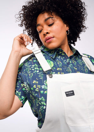 1X= A person with curly hair, wearing a floral shirt and The Essential Herringbone Overall in light colors with adjustable straps, poses on a plain white background with eyes closed and hand on the strap. The overalls feature a rectangular black patch adorned with a logo.