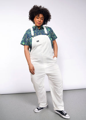 1X= A person with curly hair poses stylishly in a studio, wearing a blue floral short-sleeve shirt and The Essential Herringbone Overall with adjustable straps. Their hand rests casually in the deep pocket of the overalls, complemented by black sneakers against a gray background.