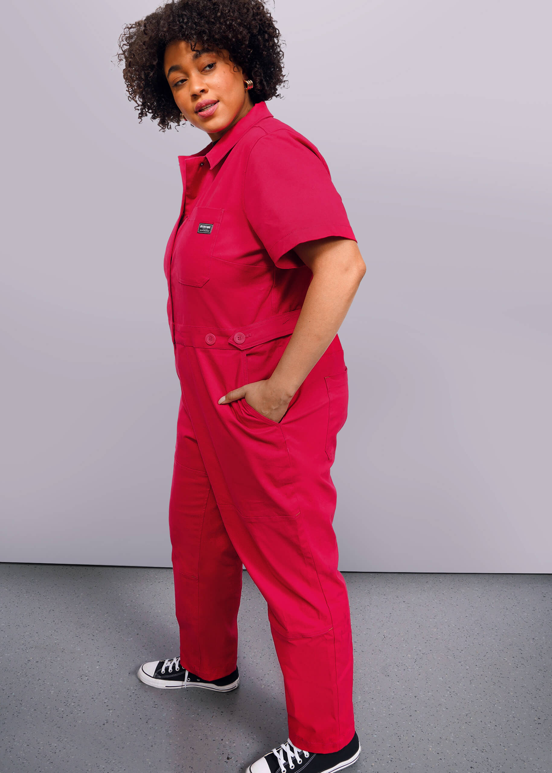1X=A person with curly hair wearing The Essential High Waisted Coverall in bright pink with deep pockets and black sneakers stands against a plain white background, hands in pockets, gazing confidently to the side.