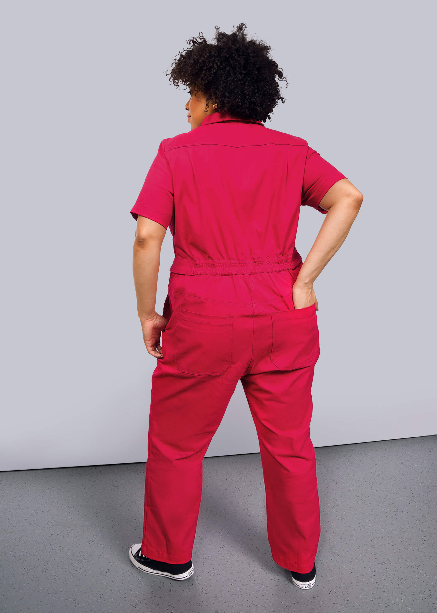 1X=On a plain white background, a curly-haired individual wears The Essential High Waisted Coverall in bright pink paired with black sneakers, its adjustable waist and high-waisted fit elegantly highlighting their silhouette as they confidently face away on the light gray floor.