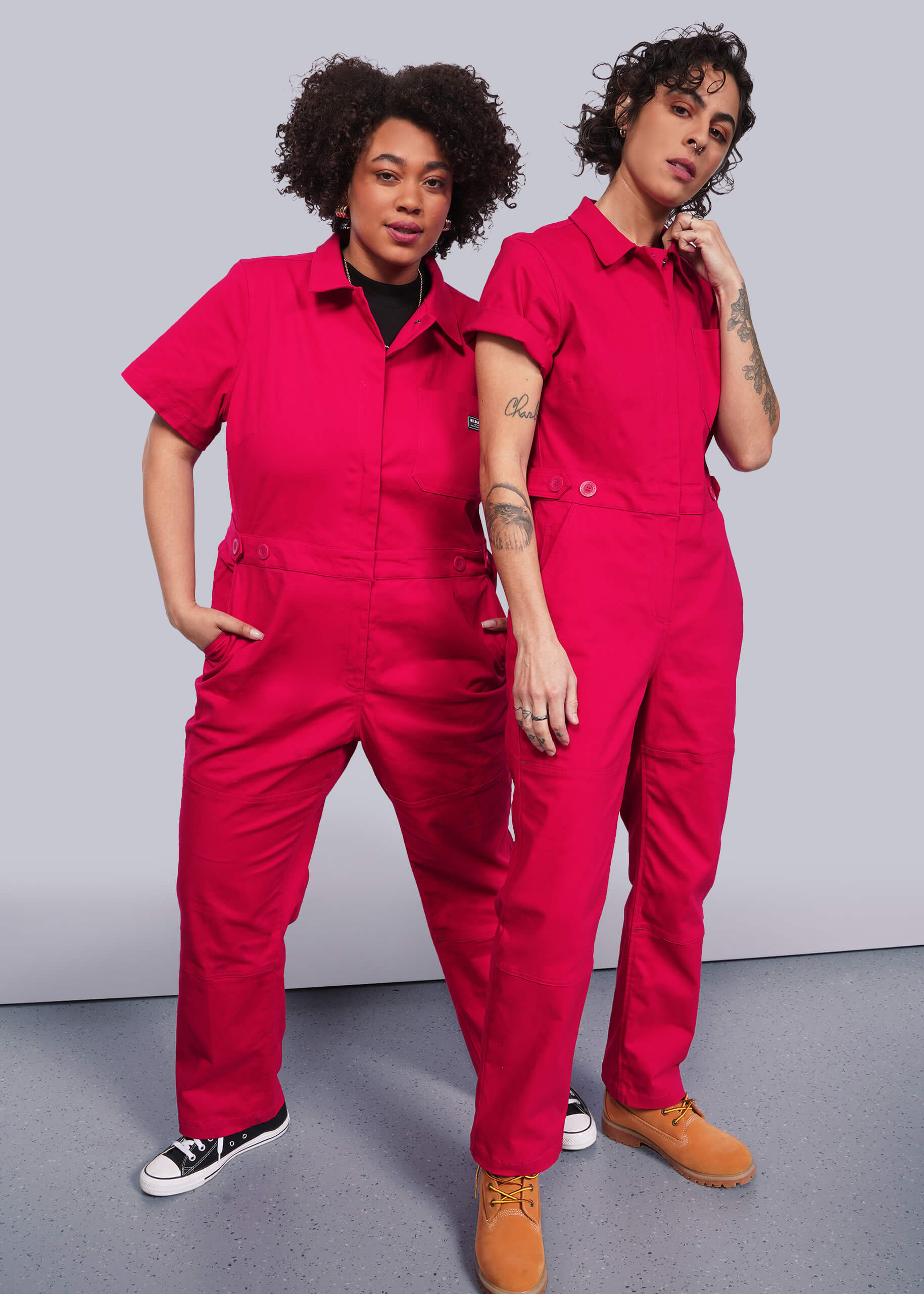 Two people confidently wear bright pink The Essential High Waisted Coverall. One, with hands in deep pockets, pairs it with sneakers, while the other stands thoughtfully with boots. Both have curly hair and visible tattoos against a plain backdrop.