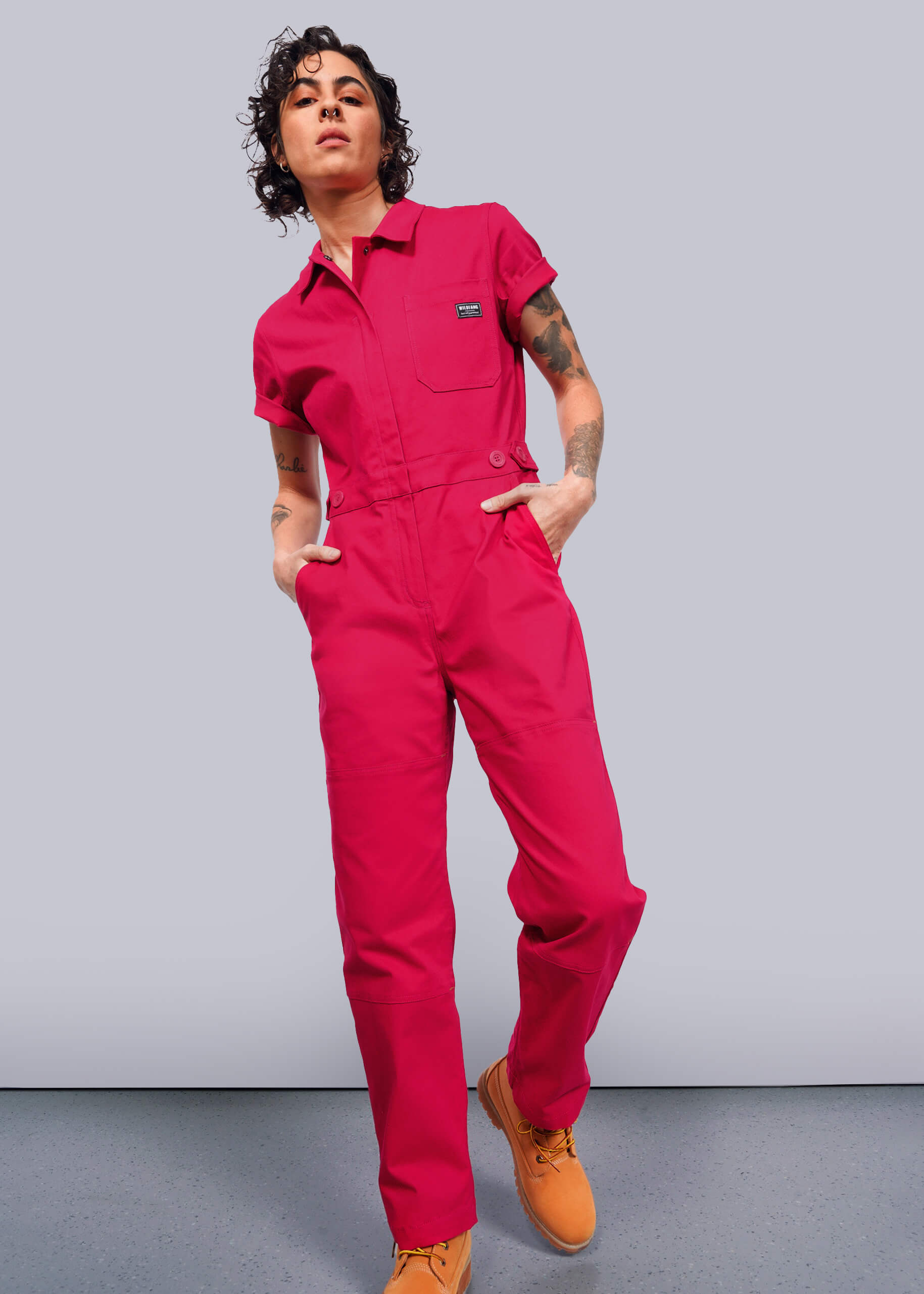 S=A person with curly hair confidently stands in The Essential High Waisted Coverall, a bright pink jumpsuit with an adjustable waist and deep pockets, paired with tan work boots. Against a white background, they have tattoos on their arms and hands in pockets, exuding style with a slight lean and head tilt.