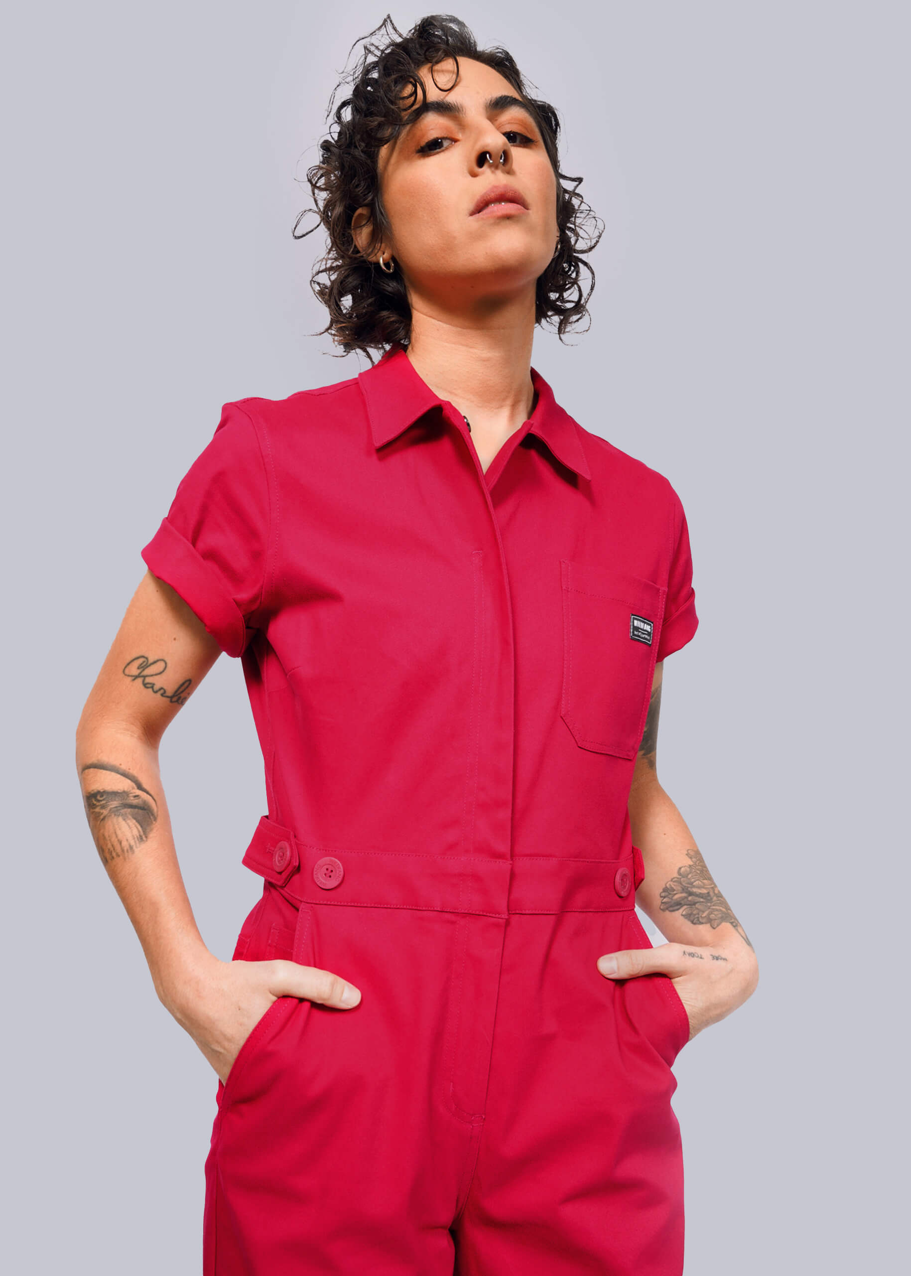 S=Wearing The Essential High Waisted Coverall in bright red, a person stands confidently against a white background. With curly hair and visible tattoos, they have the sleeves rolled up and their hands in deep pockets.