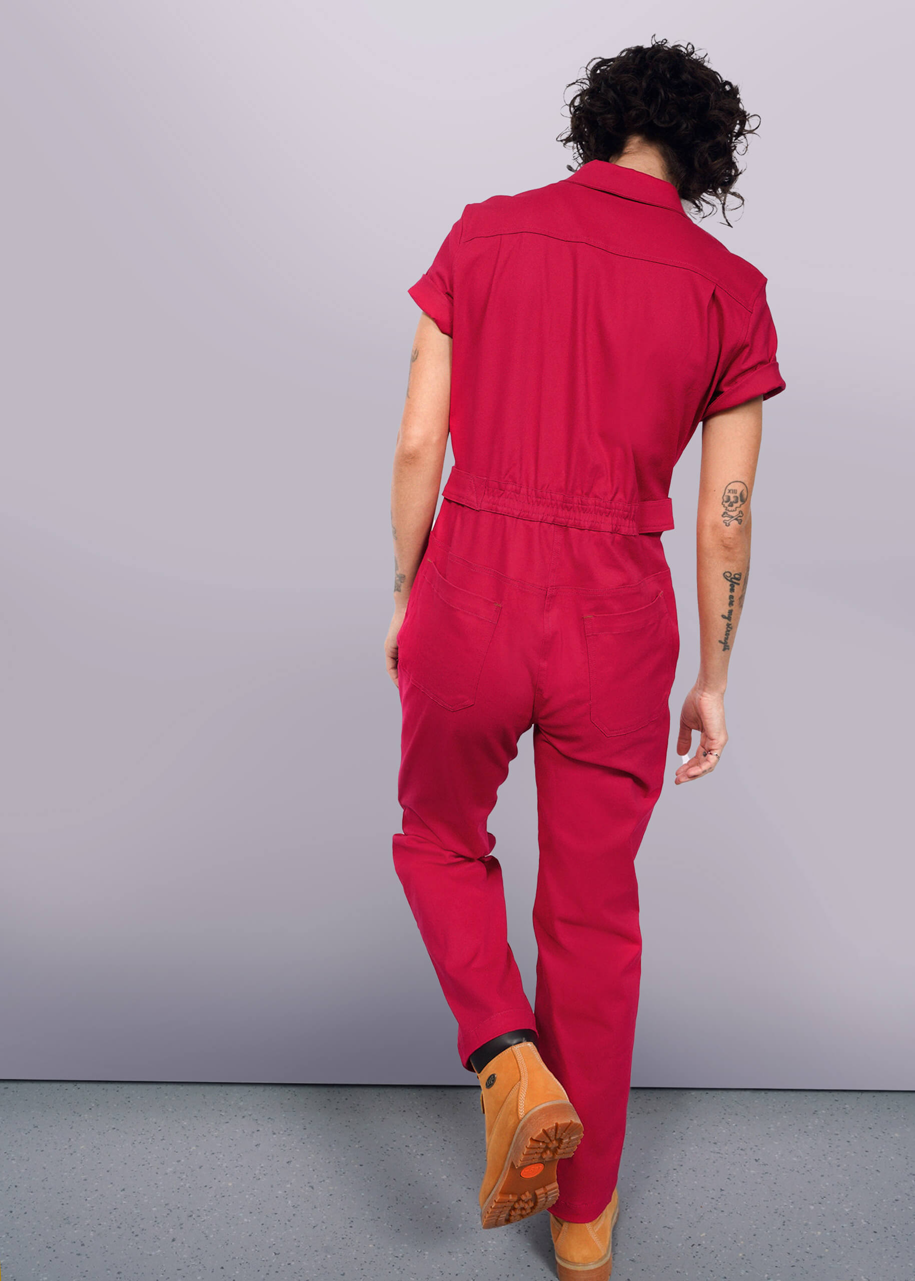 S=In a vibrant red Essential High Waisted Coverall with adjustable waist and deep pockets, a person with curly hair walks away from the camera. Their left arm tattoos are visible through short sleeves as they stride confidently in tan boots against a plain white background.