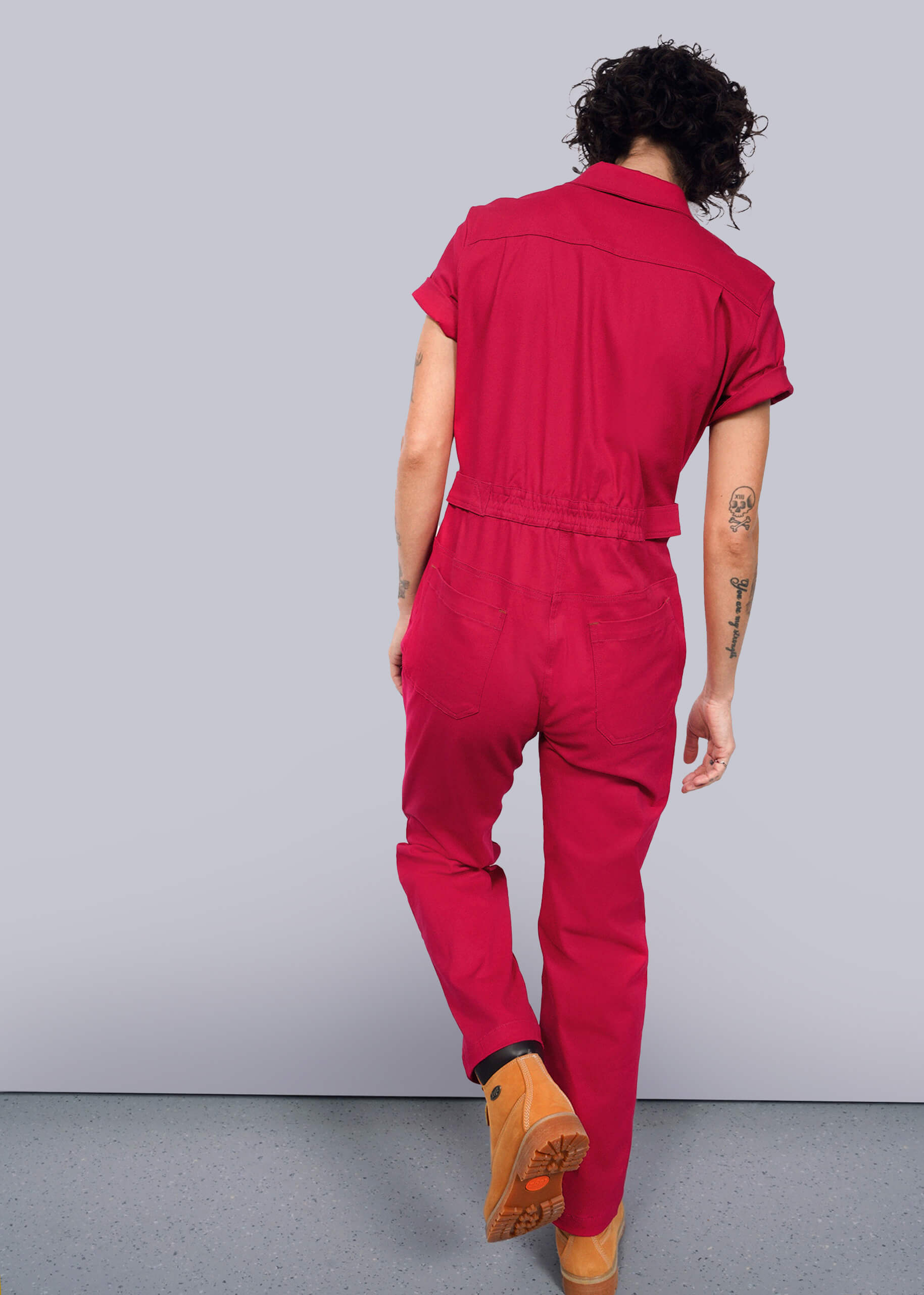 S=In a vibrant red Essential High Waisted Coverall with adjustable waist and deep pockets, a person with curly hair walks away from the camera. Their left arm tattoos are visible through short sleeves as they stride confidently in tan boots against a plain white background.