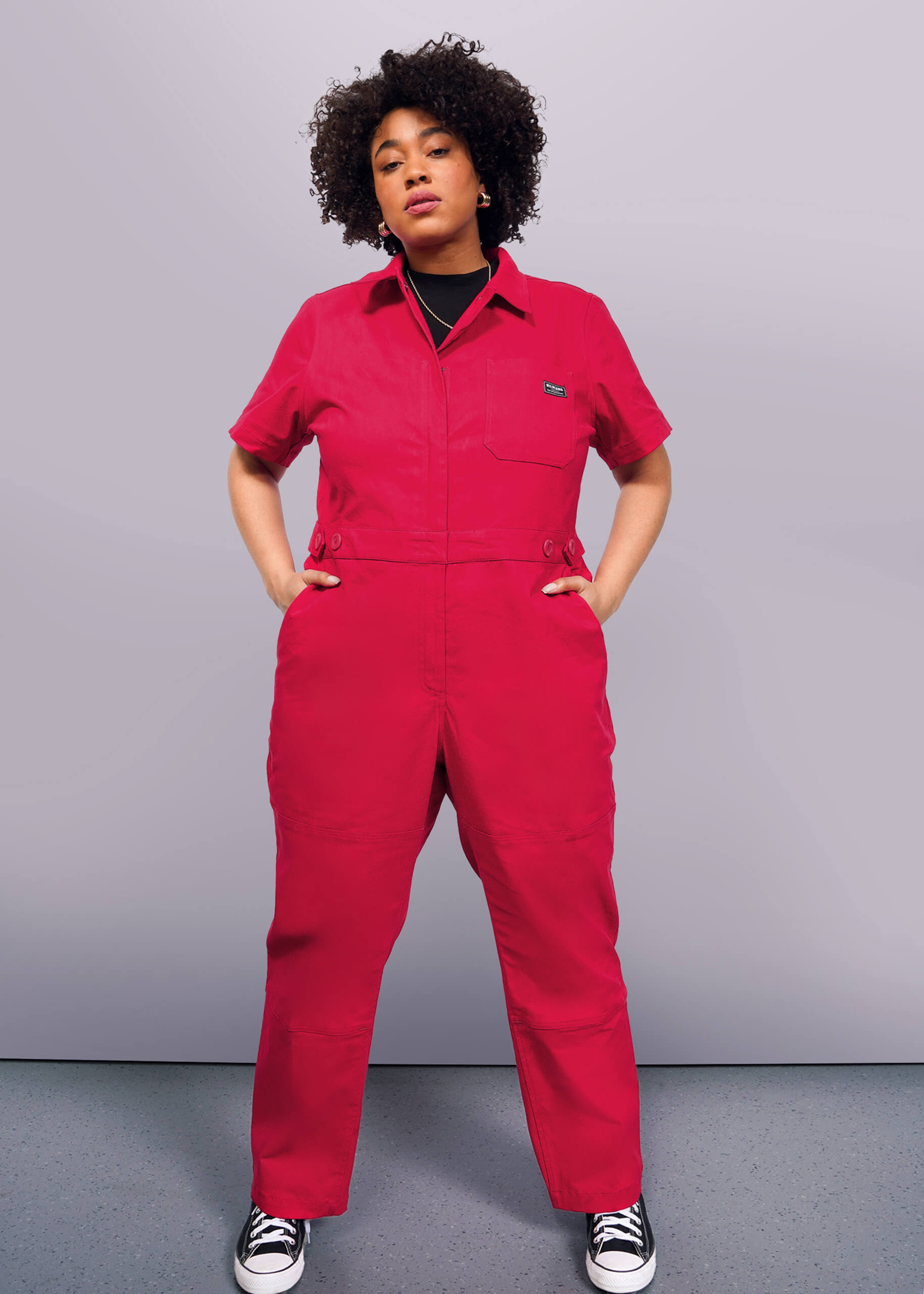 1X=A person poses confidently against a plain backdrop, styled in The Essential High Waisted Coverall—a bright red jumpsuit with an adjustable waist—paired with black-and-white sneakers. Their hands rest in deep pockets, and they sport curly hair.