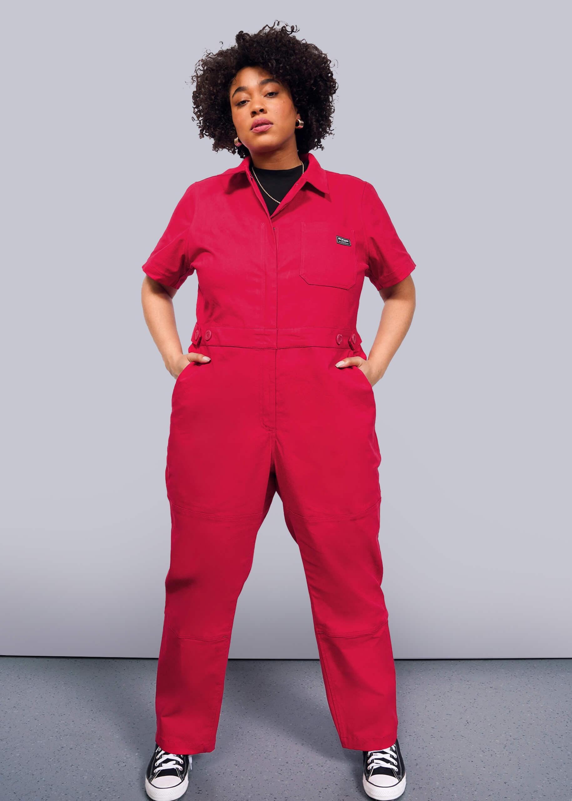 1X=A person poses confidently against a plain backdrop, styled in The Essential High Waisted Coverall—a bright red jumpsuit with an adjustable waist—paired with black-and-white sneakers. Their hands rest in deep pockets, and they sport curly hair.