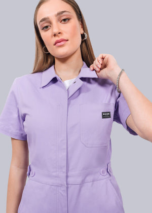 S= A woman stands confidently against a white background, wearing The Essential High Waisted Coverall in light purple cotton stretch with short sleeves, front pocket, and adjustable waist, holding the collar with one hand.