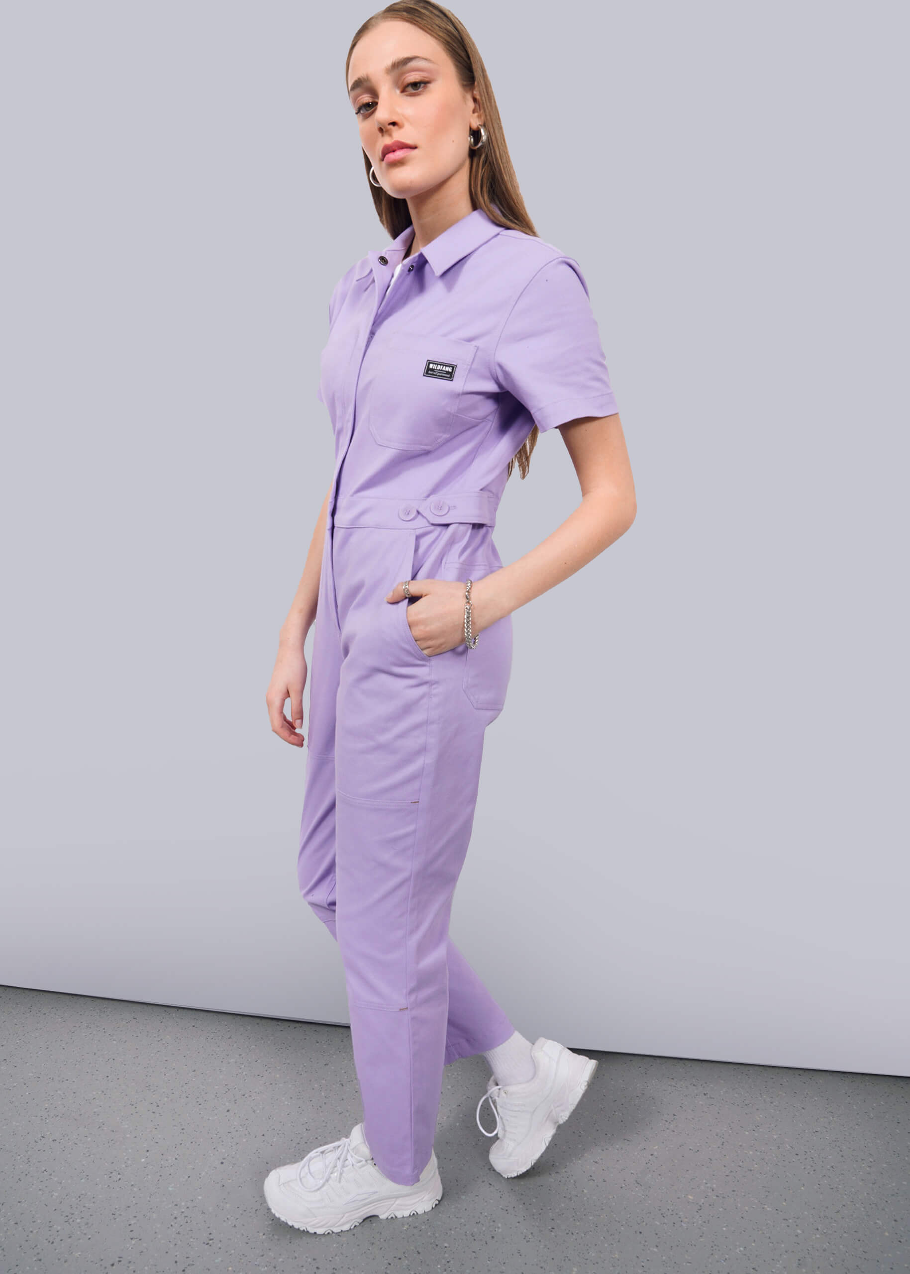 S=A woman stands sideways against a white wall, wearing The Essential High Waisted Coverall in light purple with short sleeves and an adjustable waist. Her hands are in her pockets as she confidently poses, styled with white sneakers.