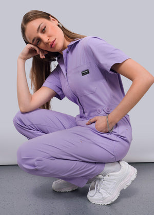 S=A woman wearing The Essential High Waisted Coverall in light purple and white sneakers squats on a gray floor, resting her head on her hand and gazing at the camera with a neutral expression.