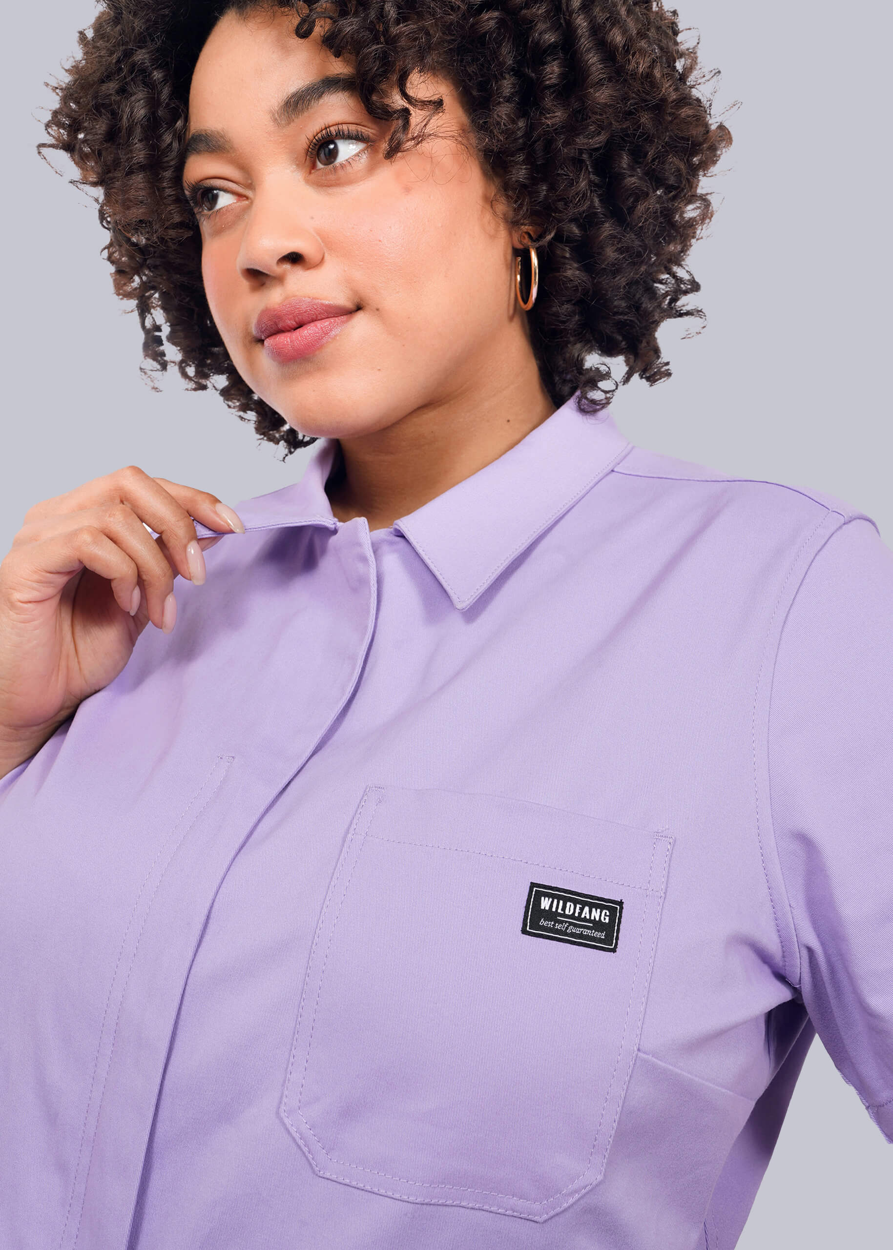 1X=A woman with curly hair wears The Essential High Waisted Coverall in light purple, featuring a collared design, front pocket with WIDIANG logo patch, and soft cotton stretch fabric, looking slightly to the side against a white background.