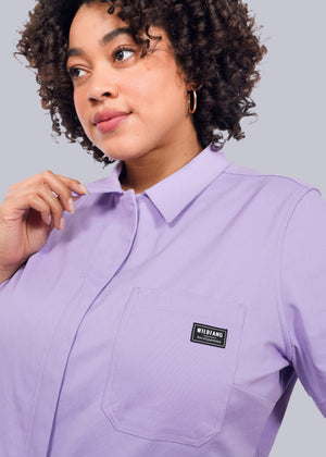 1X=A woman with curly hair wears The Essential High Waisted Coverall in light purple, featuring a collared design, front pocket with WIDIANG logo patch, and soft cotton stretch fabric, looking slightly to the side against a white background.
