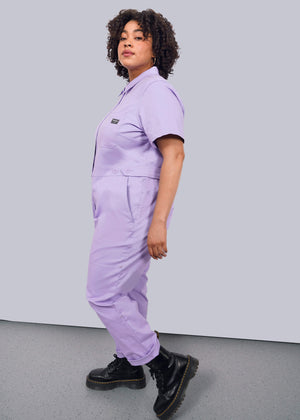 1X=A person with curly hair stands in profile against a white background, wearing The Essential High Waisted Coverall in light purple and black lace-up boots.