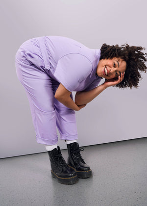 1X=A person with curly hair wears The Essential High Waisted Coverall in light purple and black boots, bending forward and smiling at the camera with one hand on their face, set against a plain white background.