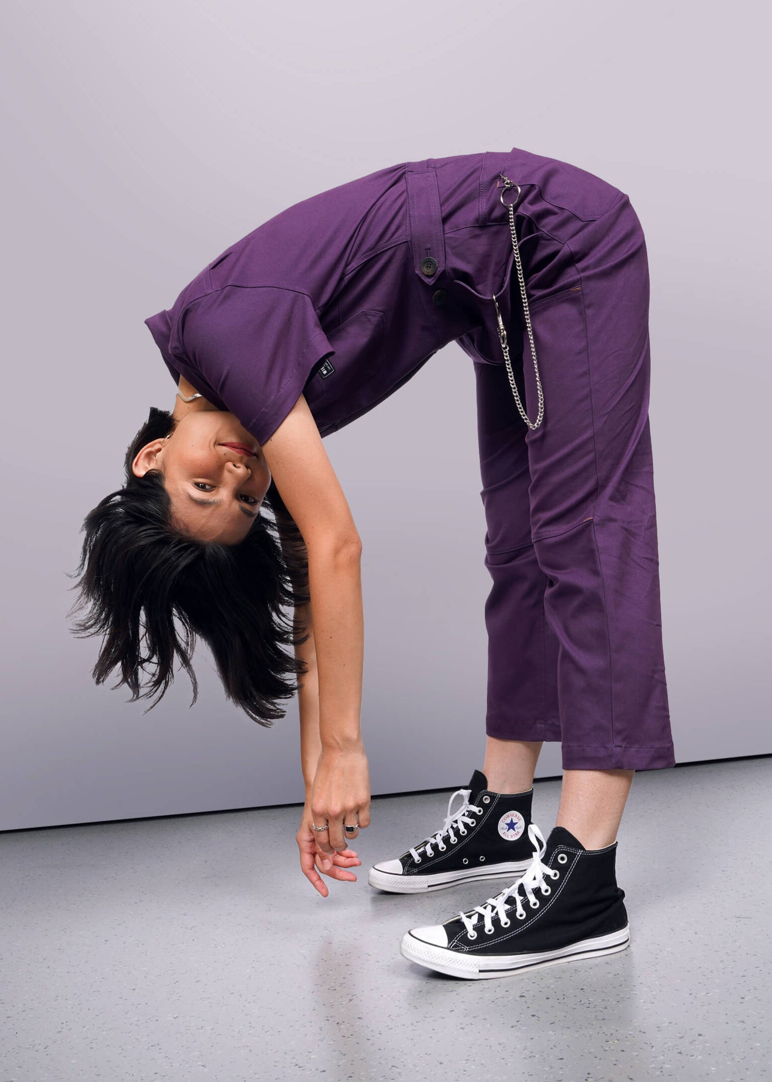 S=Person wearing a purple jumpsuit with black high-top sneakers on a gray background