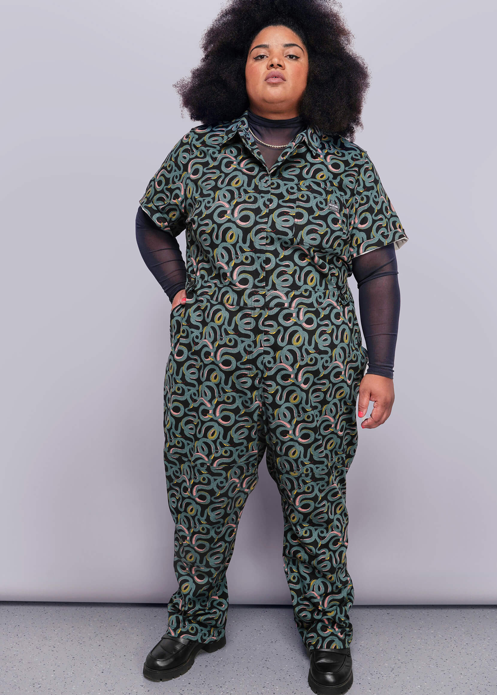 1X=Person wearing a green patterned jumpsuit against a plain background